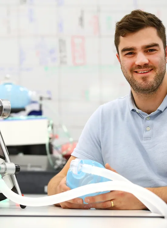 Man in a light blue shirt smiling, seated at a desk with a medical ventilation device, whiteboard in the background.