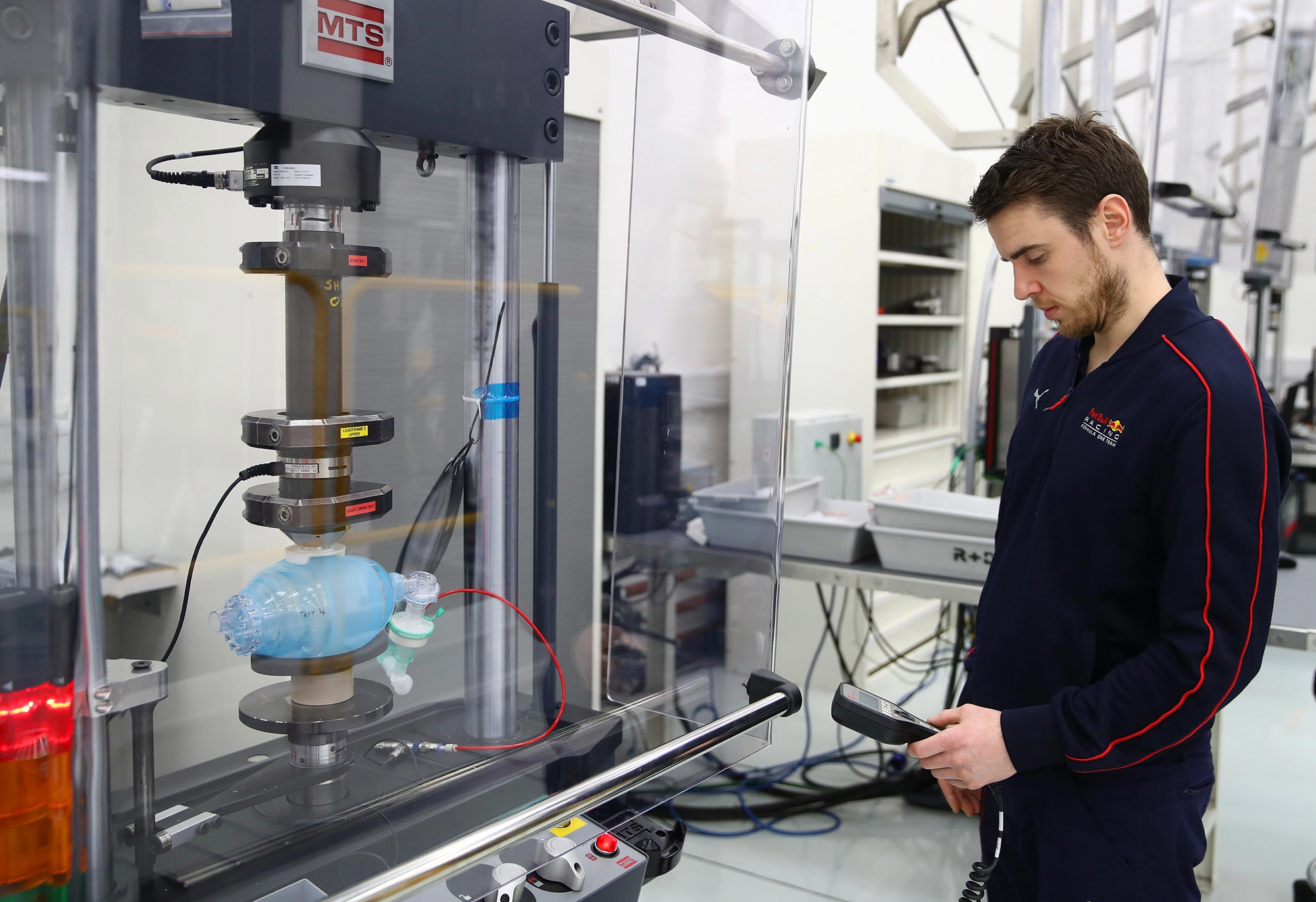 A man operates a testing device in a lab, monitoring a blue medical pump connected to wires within a protective glass enclosure.