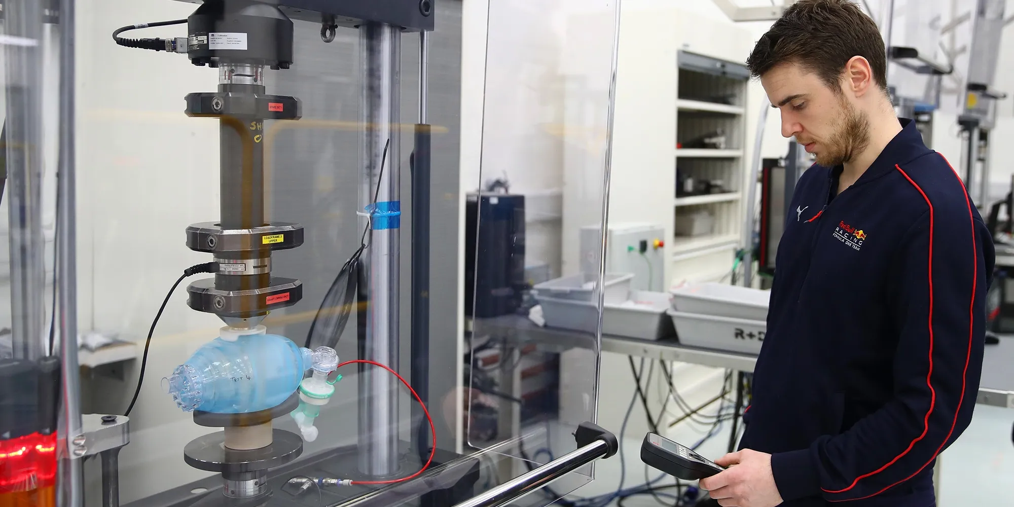 A man operates a testing device in a lab, monitoring a blue medical pump connected to wires within a protective glass enclosure.