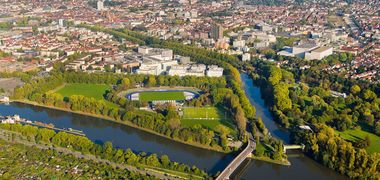 Eine Luftaufnahme von Heilbronn, eine Flussgabelung des Neckars prominent im Bild, umsäunt von grünen Bäumen.