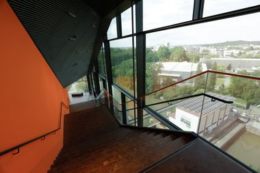 Blick eine Treppe hinunter im Gebäude der experimenta - rechter Hand schweift der Blick durch die Glasfassade hindurch über grüne Parks, ein mit Solarpaneelen bedecktes Flachdach und die Stadt im Hintergrund.