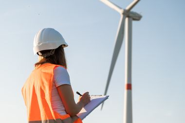 Eine Frau mit Helm und orangener Warnweste steht vor einem Windrad und notiert etwas auf einem Klemmbrett
