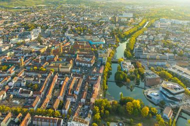 Eine Luftaufnahme von Heilbronn, rote Ziegeldächer leuchten neben grünen Baumkronen im Abendlicht. Blauer Himmel spiegelt sich im Neckar, der sich durchs Stadtgebiet schlängelt.