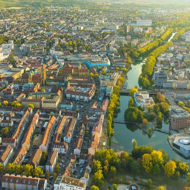 Eine Luftaufnahme von Heilbronn, rote Ziegeldächer leuchten neben grünen Baumkronen im Abendlicht. Blauer Himmel spiegelt sich im Neckar, der sich durchs Stadtgebiet schlängelt.