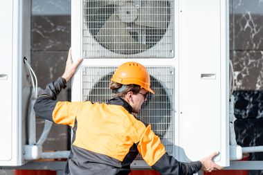 Ein Handwerker in Arbeitskleidung und Helm hält das große Außengerät einer Wärmepumpe oder Klimaanlage mit zwei Ventilatoren.