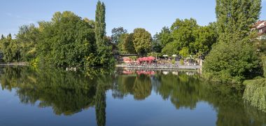 Der Weinpavillon umrahmt von grünen Bäumen an der Neckarbühne vom anderen Ufer aus gesehen. Im Vordergrund spiegelt sich die Szenerie im Neckar.