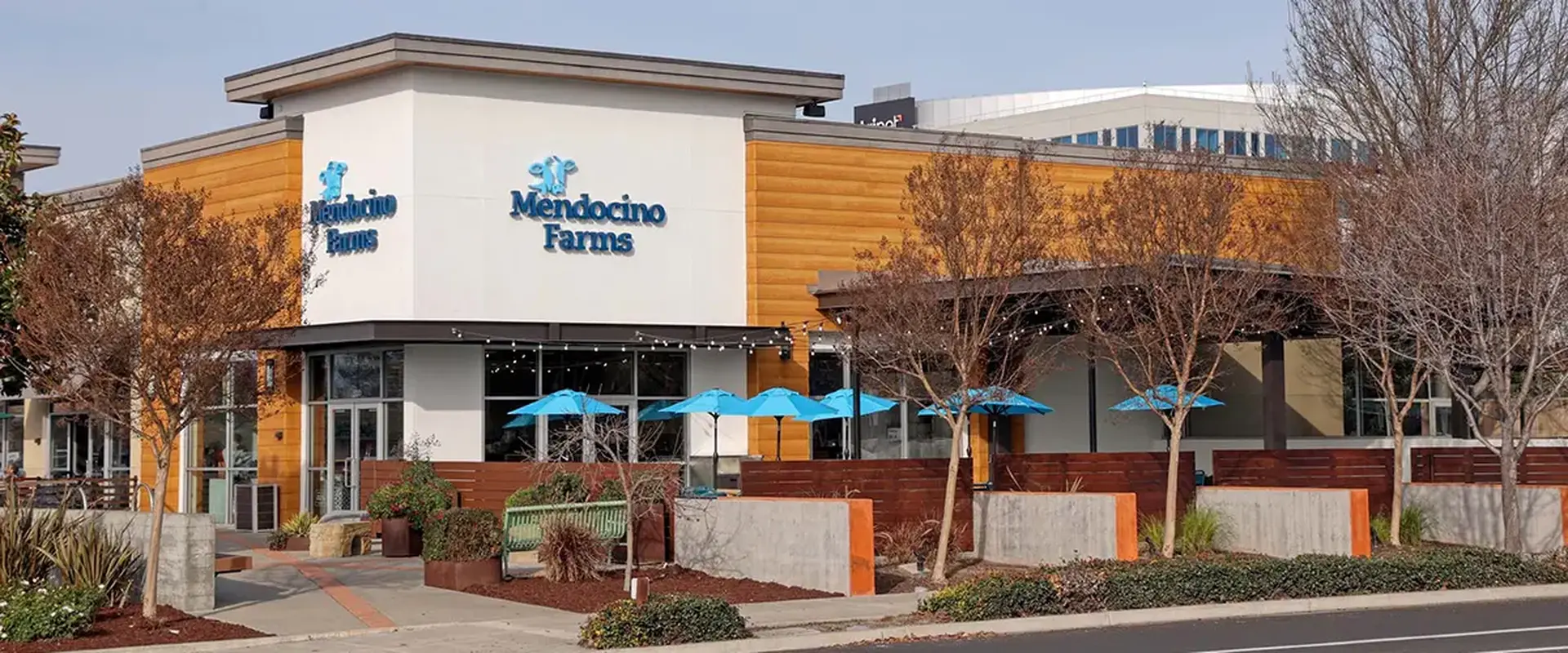 Modern Mendocino Farms exterior with warm wood accents, patio seating with blue umbrellas, and landscaped walkways.