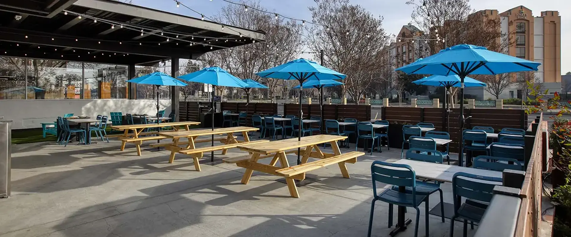Spacious outdoor patio featuring communal wooden picnic tables, bright blue umbrellas for shade, and string lighting that creates a relaxed, social dining atmosphere.