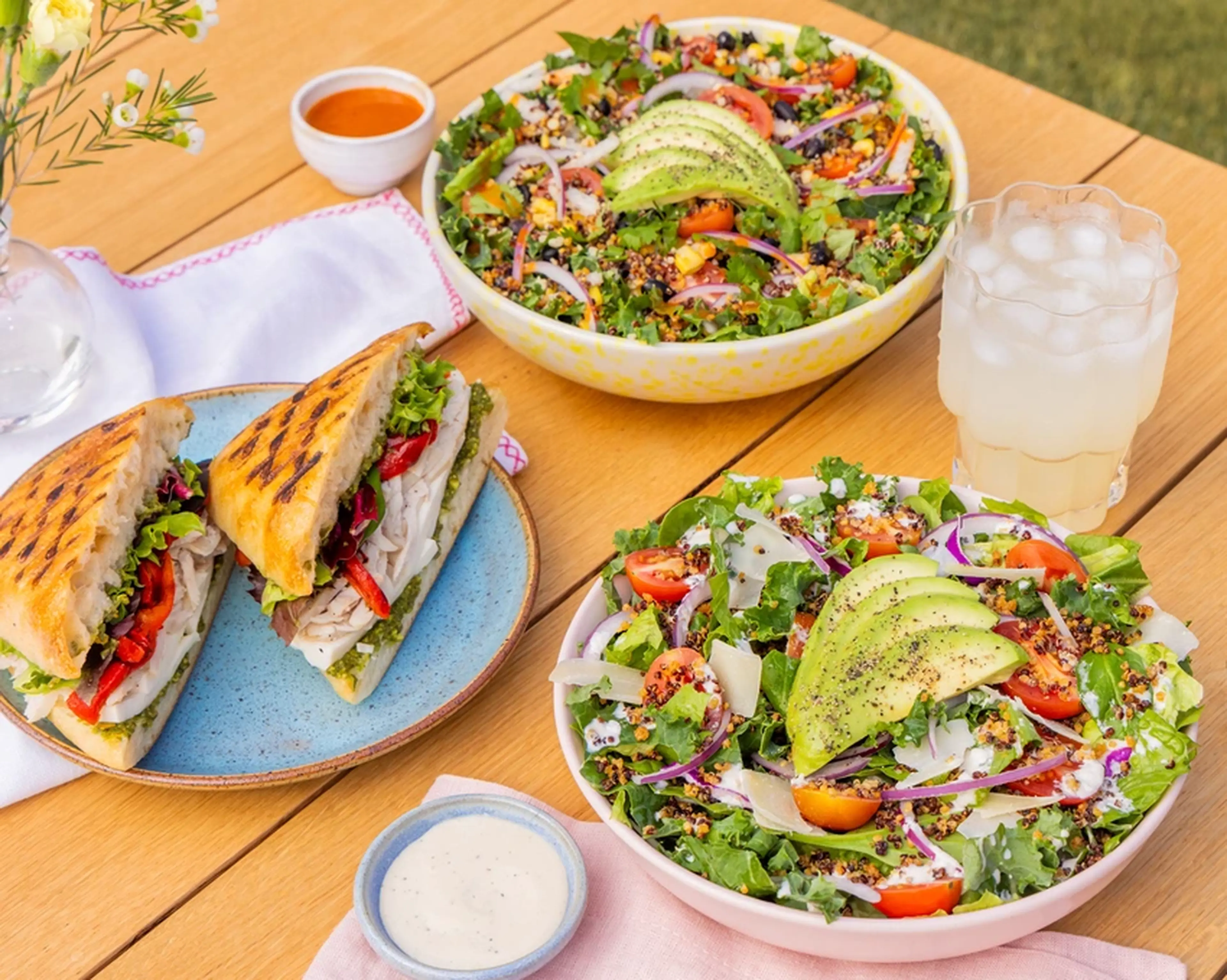 Two fresh salads with avocado, cherry tomatoes, and greens, a grilled sandwich, dressing, and a glass of iced lemonade on a wooden table.