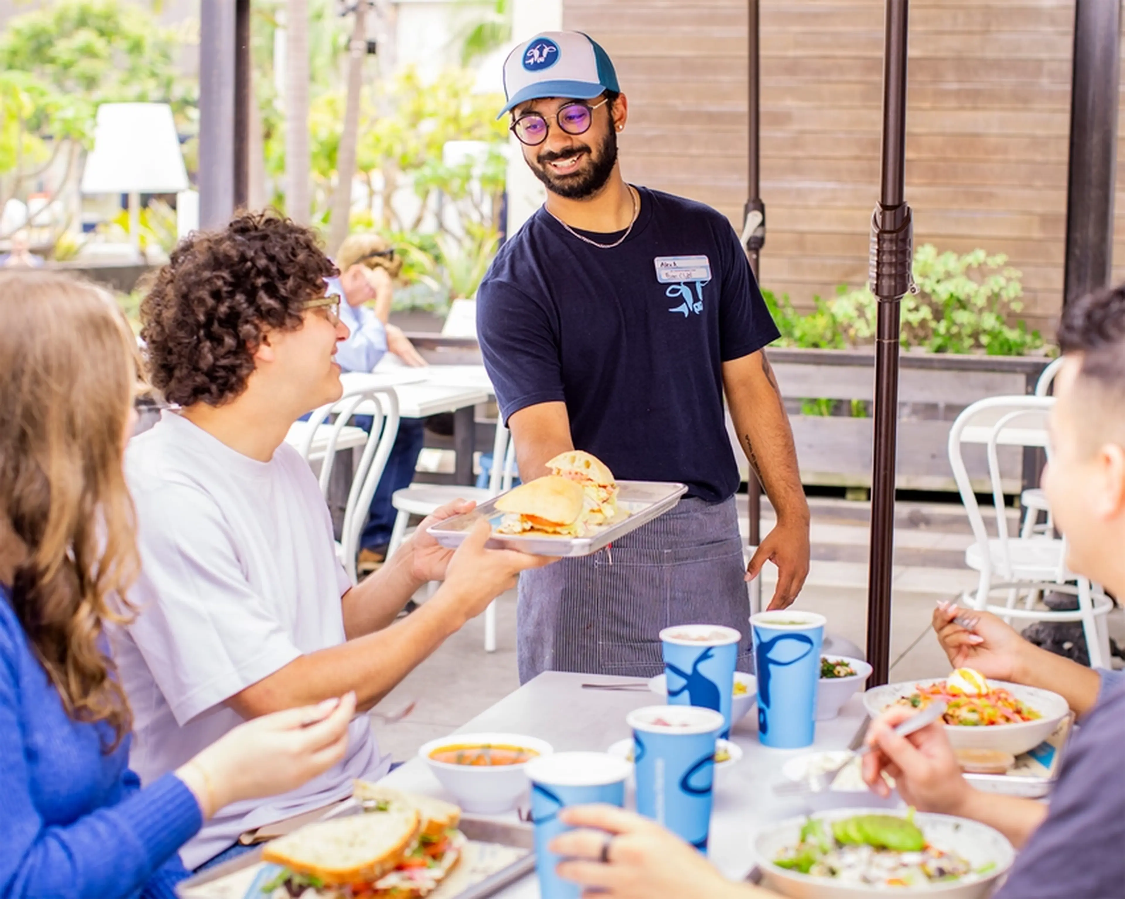 A server delivers a sandwich to a smiling group of four friends dining outdoors, with drinks and salads on the table.