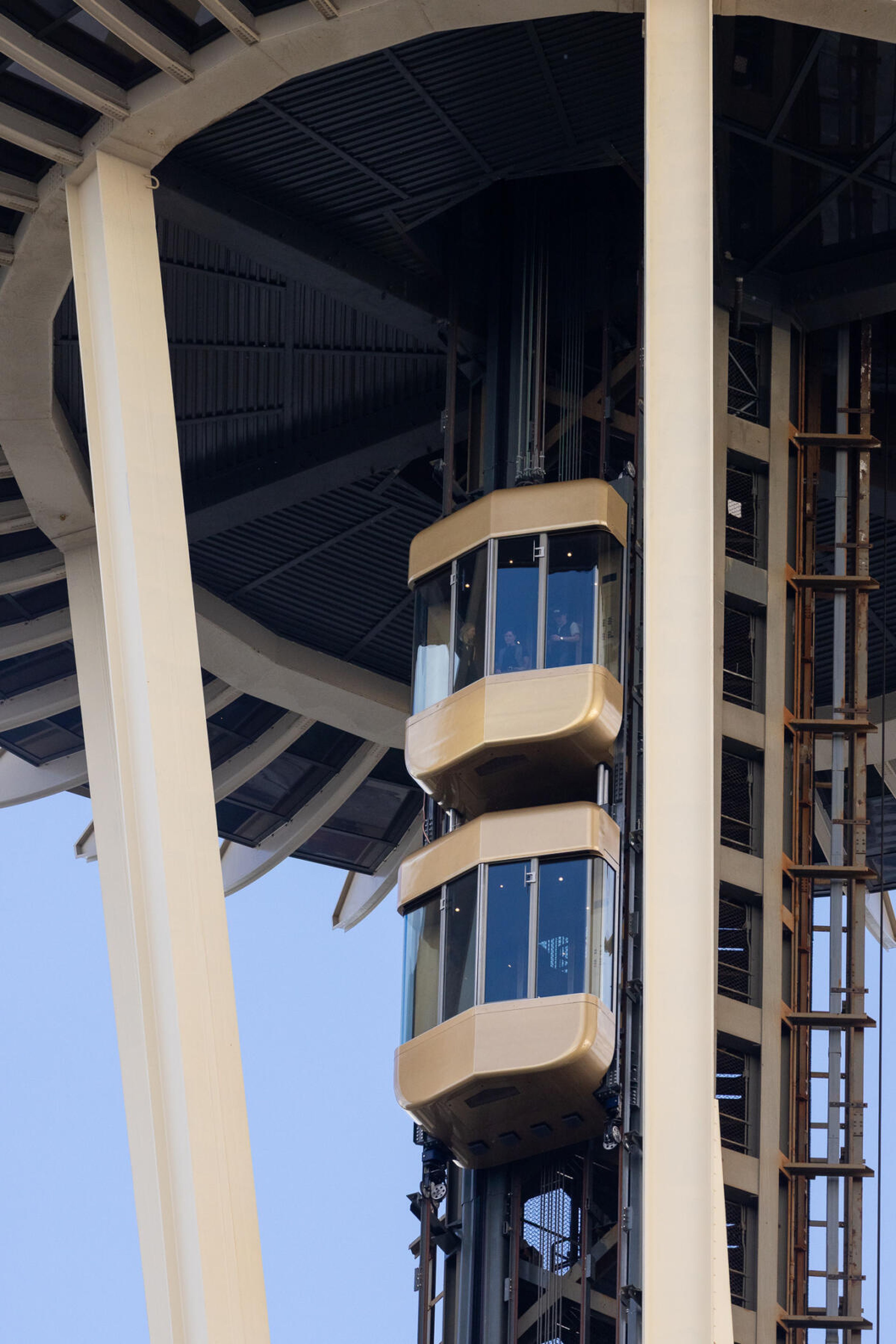 Double-height glass elevators ascending the exterior of a tall structure, with a view of the sky and metal beams.