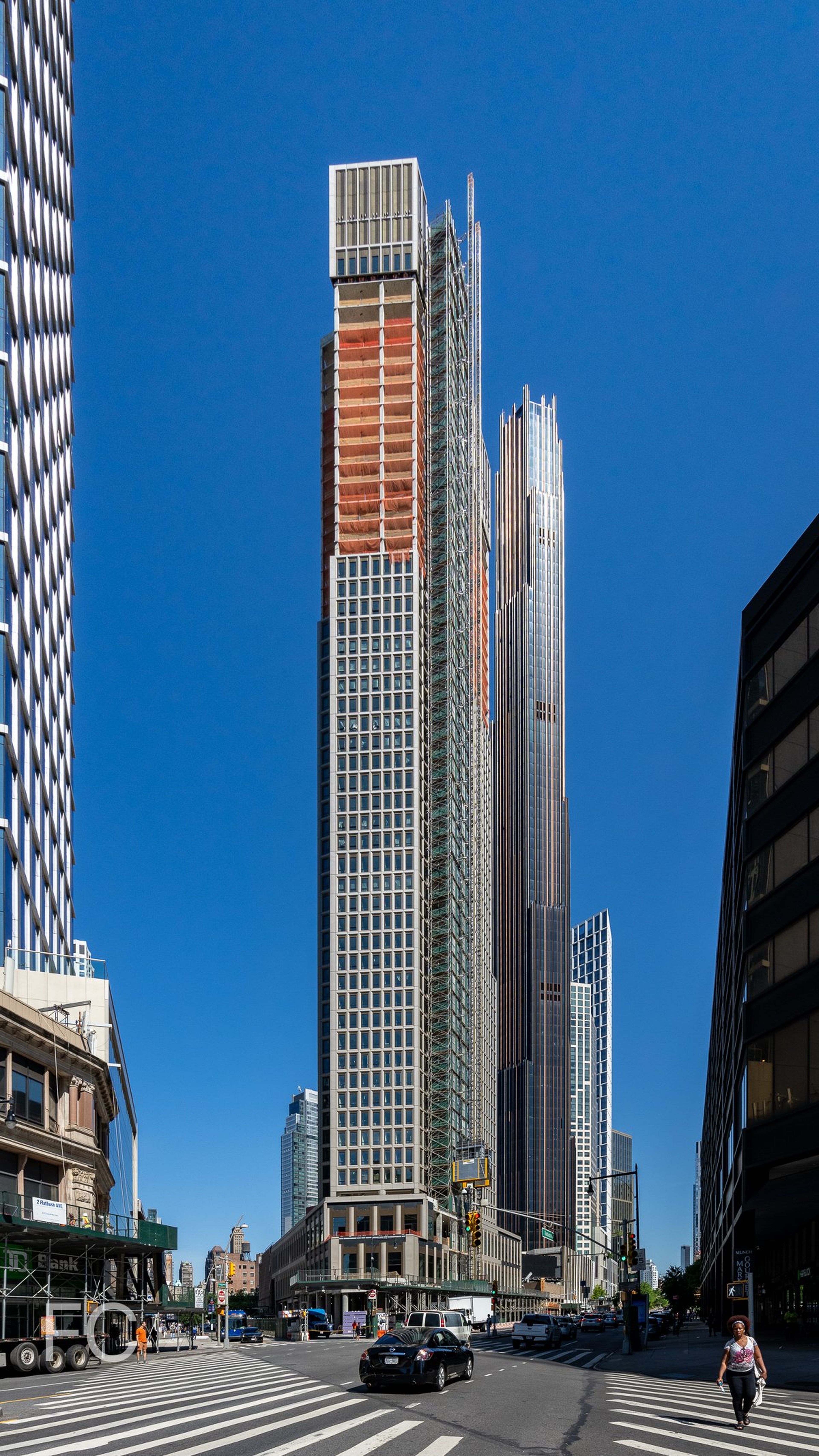 Tall skyscraper under construction with scaffolding, set against a clear blue sky, flanked by other modern buildings and a busy street below. Located at a triangular intersection.