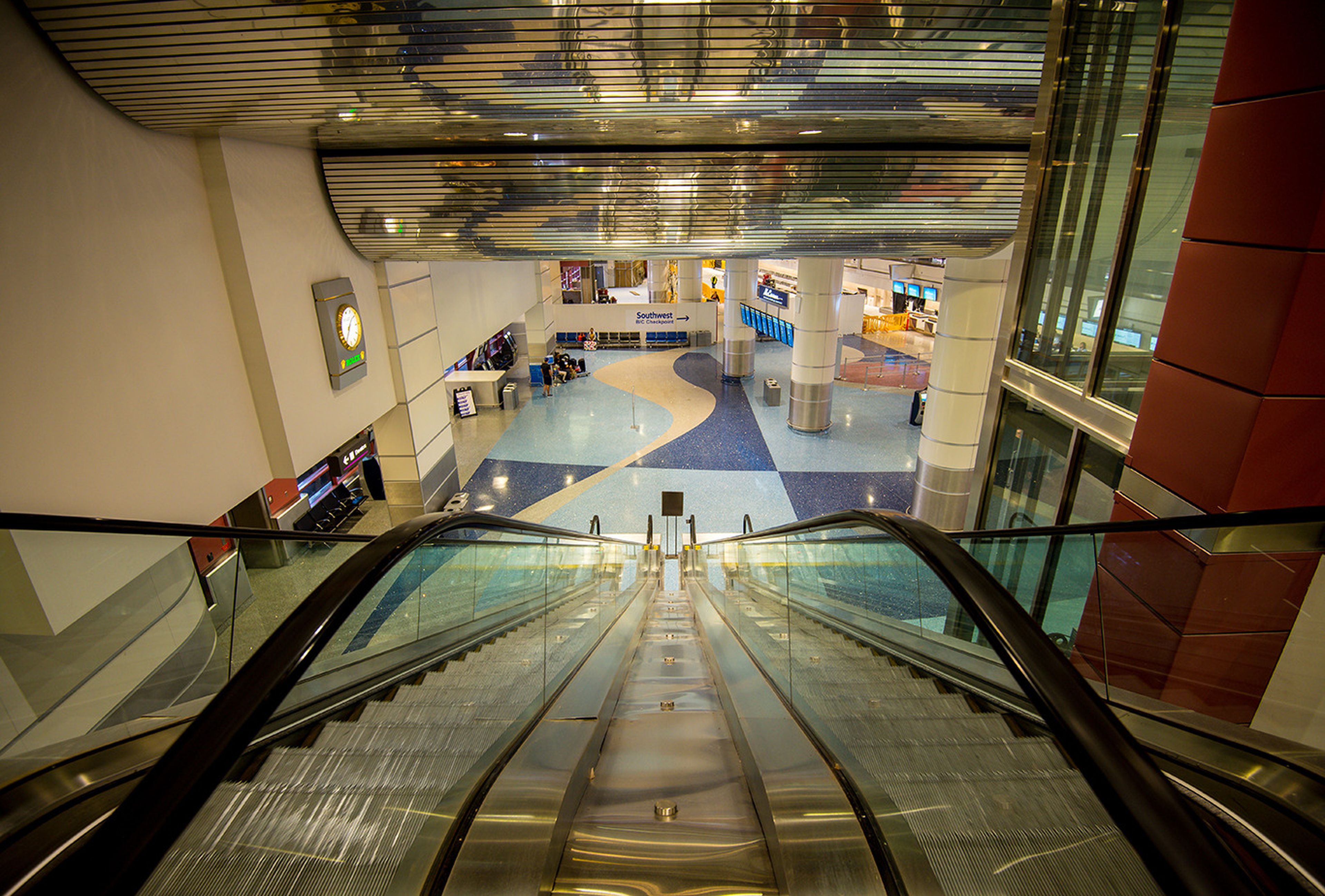 View from the top of an escalator in a modern airport terminal, featuring shiny floors, curved reflective ceiling, and a distant check-in area.