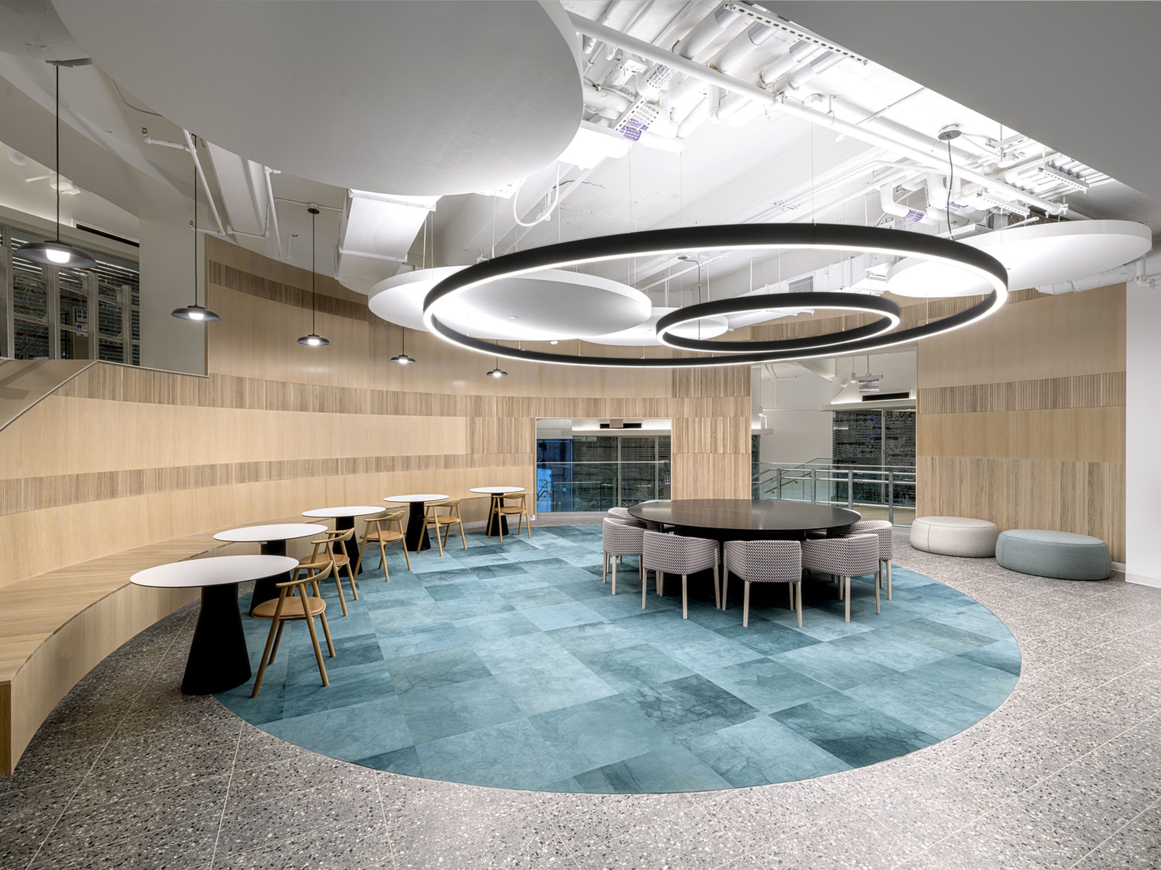 Modern office lounge with wood paneled walls and semi-circular seating banquette. The floor features blue stone tile in a circle, surrounded by gray terrazzo. In the center is a round table surrounded by upholstered chairs and large circular ceiling lights above. Two round seating pods line the other wall to the right.