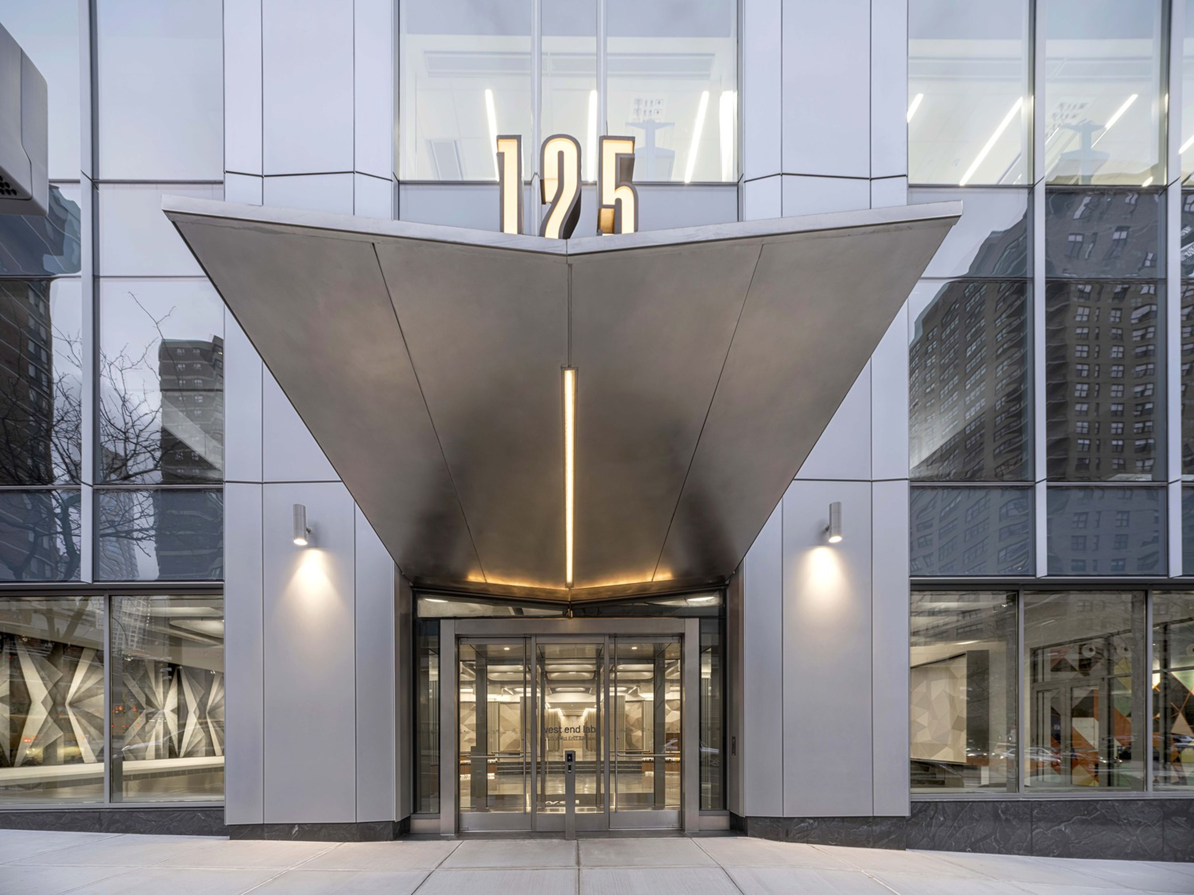 Modern building entrance with an inverted V-shaped canopy and the number "125" above the doors. Reflections of city buildings visible in the glass façade.