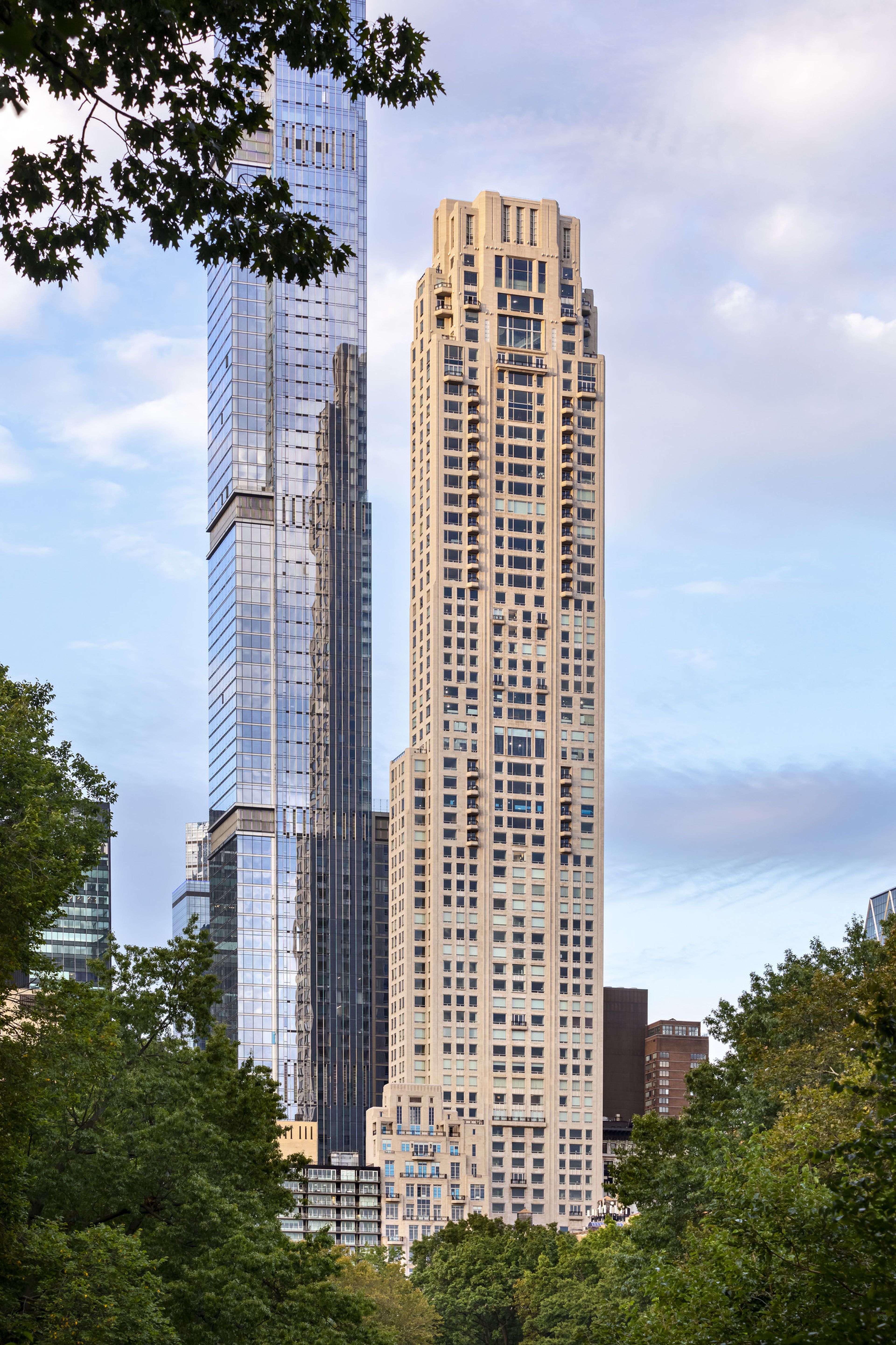 Two tall skyscrapers--one glass, one stone with rectangular windows--rise against a partly cloudy sky, framed by lush green trees at the bottom.