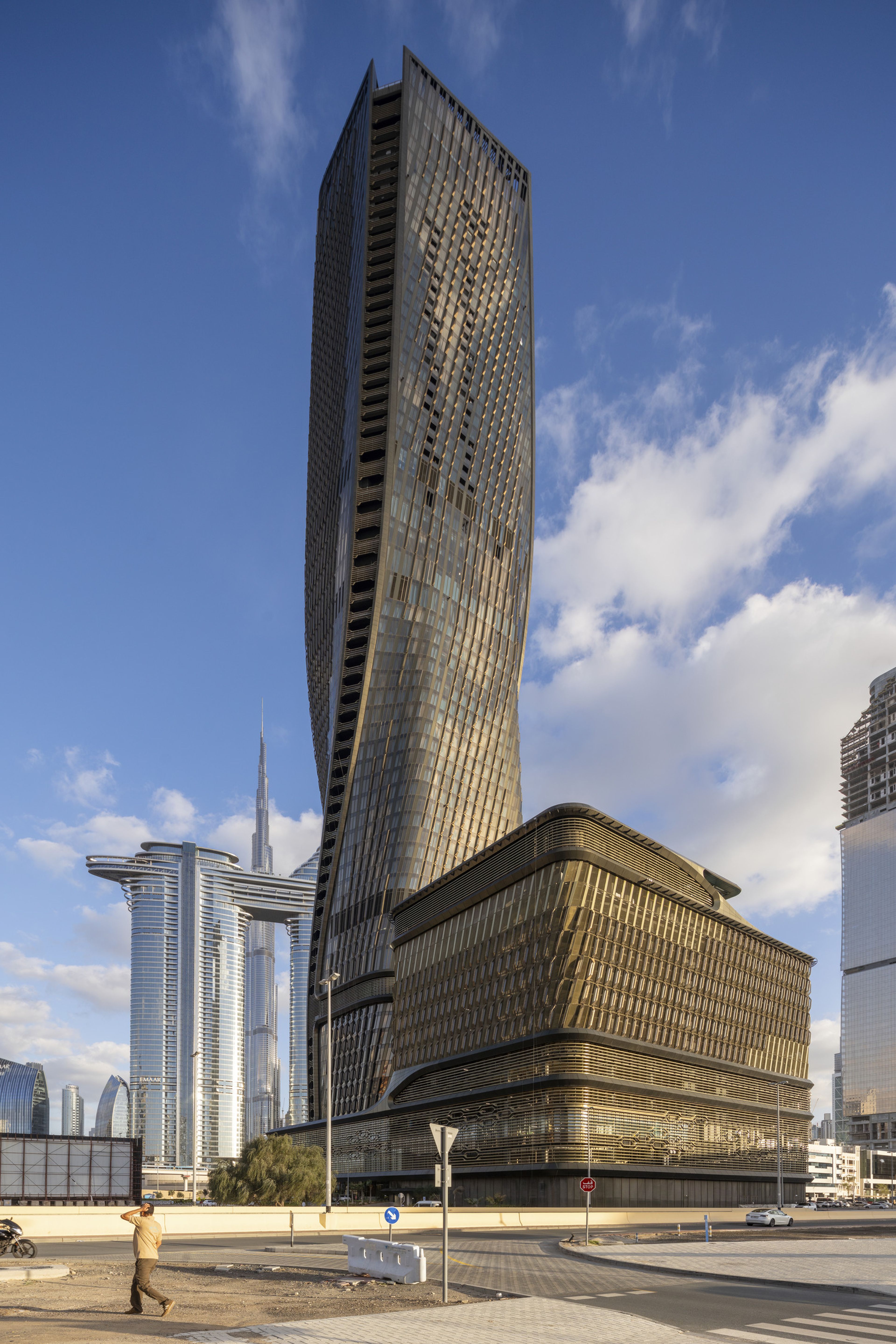 A tall, modern skyscraper with a twisting facade stands against a blue sky with scattered clouds; smaller buildings are visible in the background.