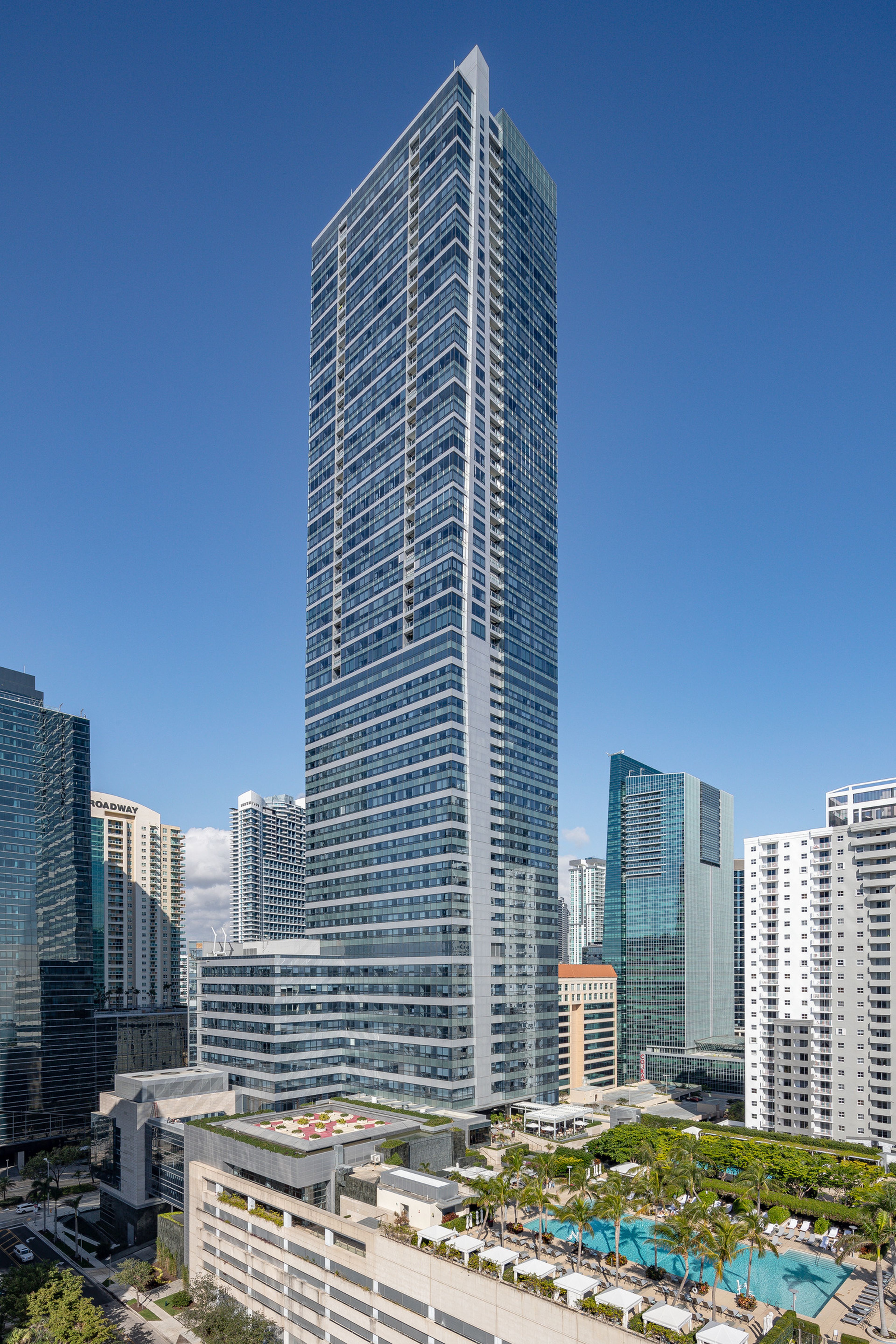 Tall modern skyscraper with glass facade with horizontal banding under a clear blue sky, surrounded by other buildings and featuring a landscaped pool deck atop a parking garage in the foreground.