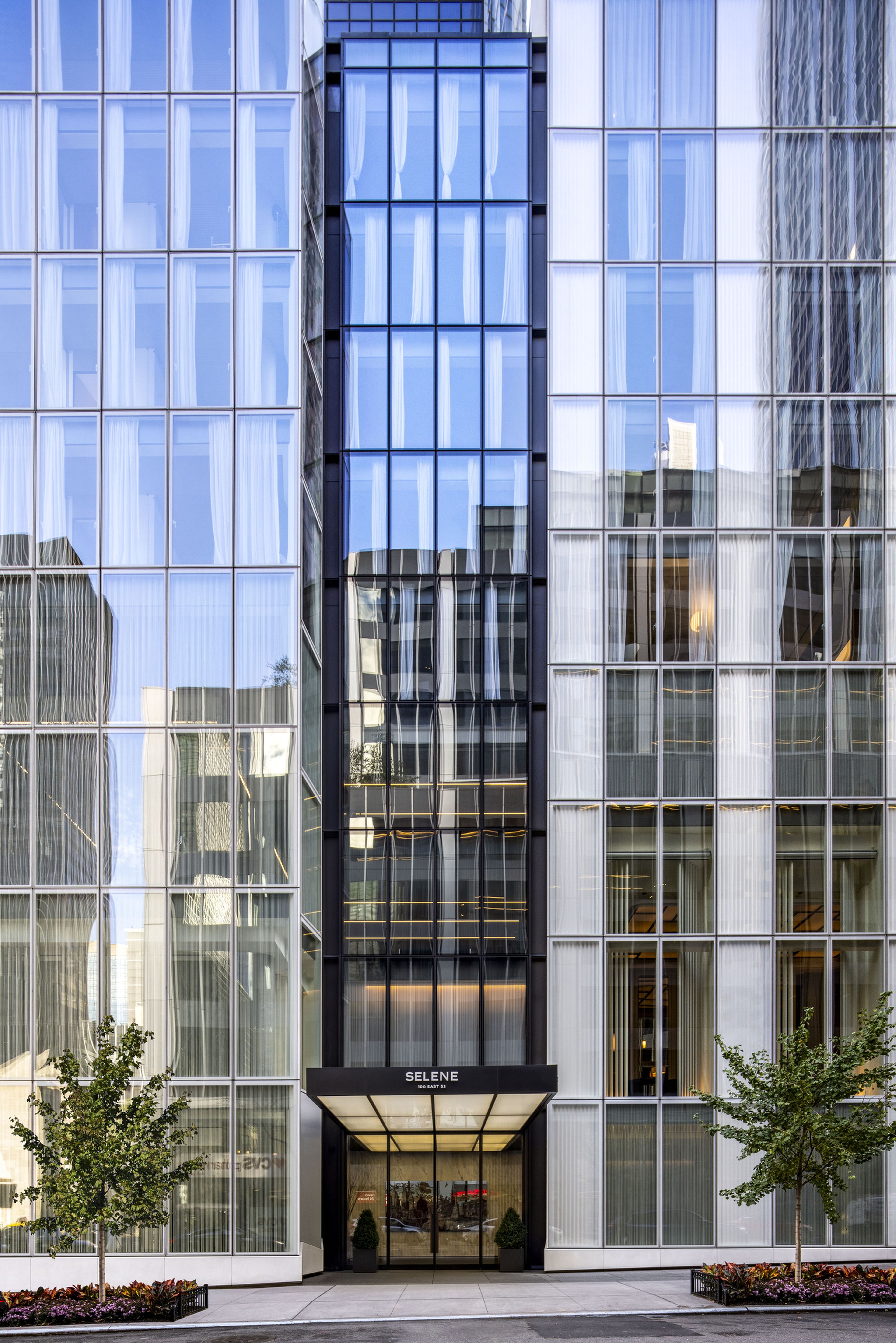 Modern glass building facade with a central entrance labeled "SELENE," flanked by two small trees and reflecting nearby structures.