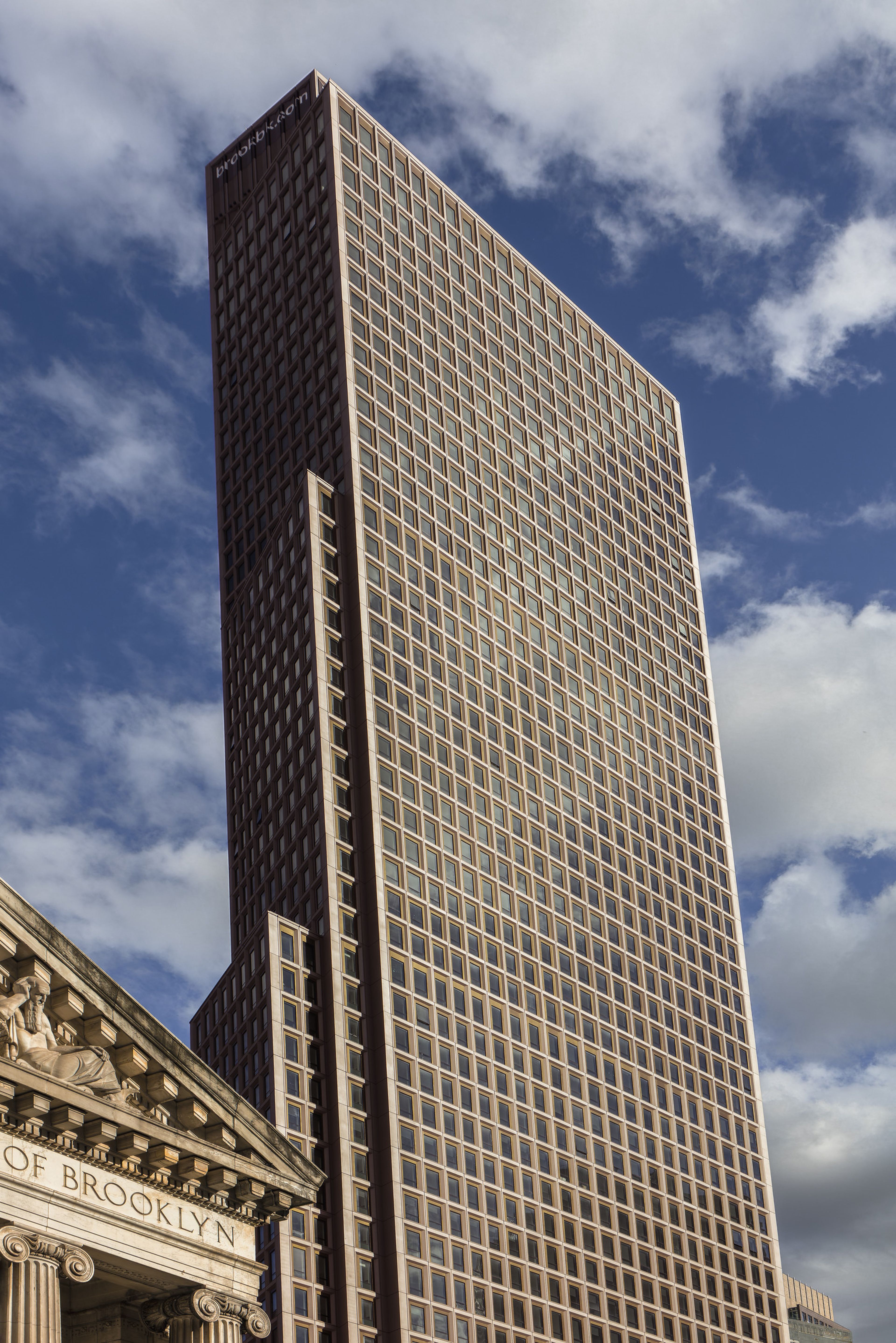 Tall, modern building with a grid of square, bronze and white framed windows set against a blue sky with white puffy clouds. The building features a banner reading "Brookbk.com" at the top. To the left is a classical style building with the words "Of Brooklyn" carved in its stone entablature.