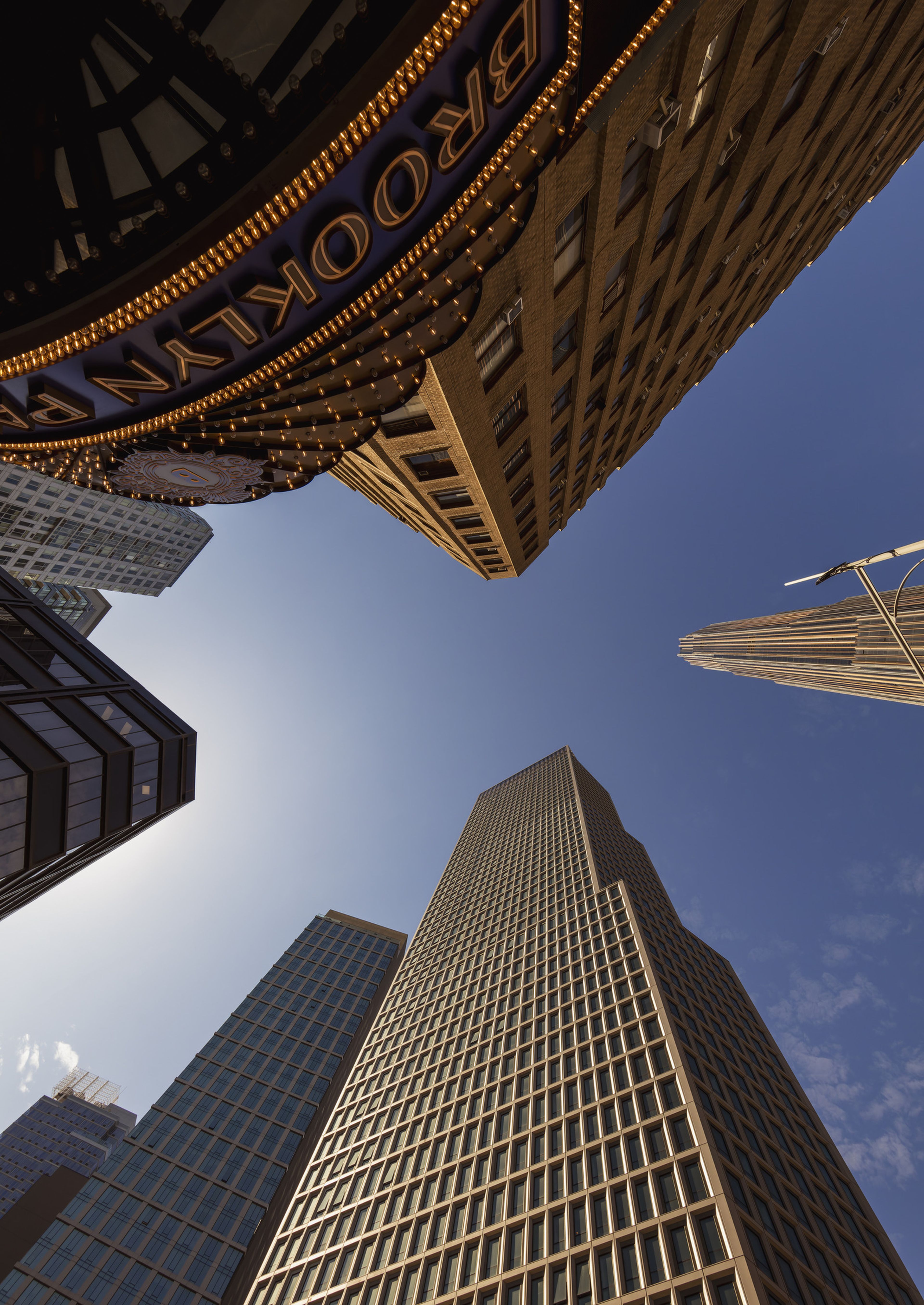Worm's eye view looking up at several tall buildings, one with a theater marquee, all in vertical perspective towards the blue sky above.
