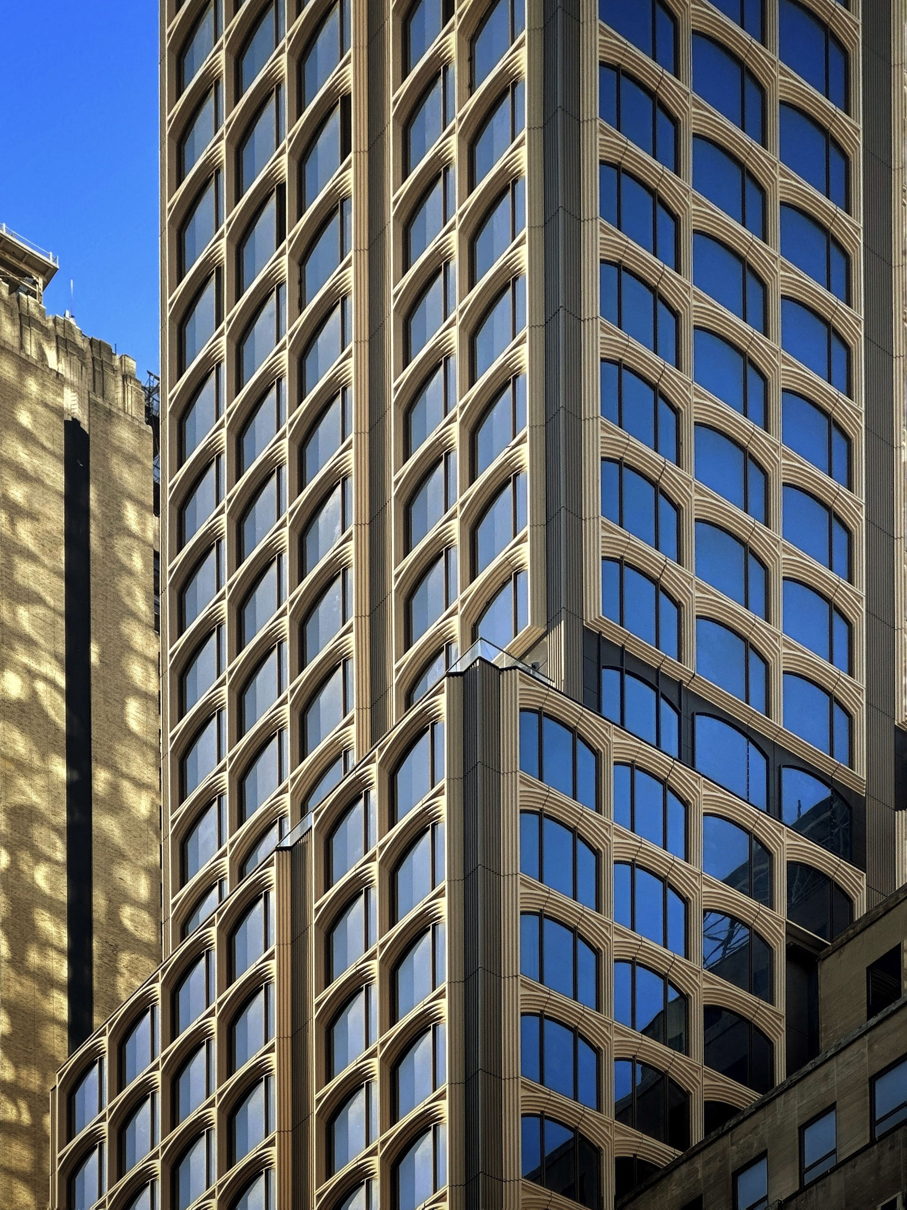 Tall building with reflective blue windows and grid-like facade against a clear blue sky. Another building's shadows are cast on its side.