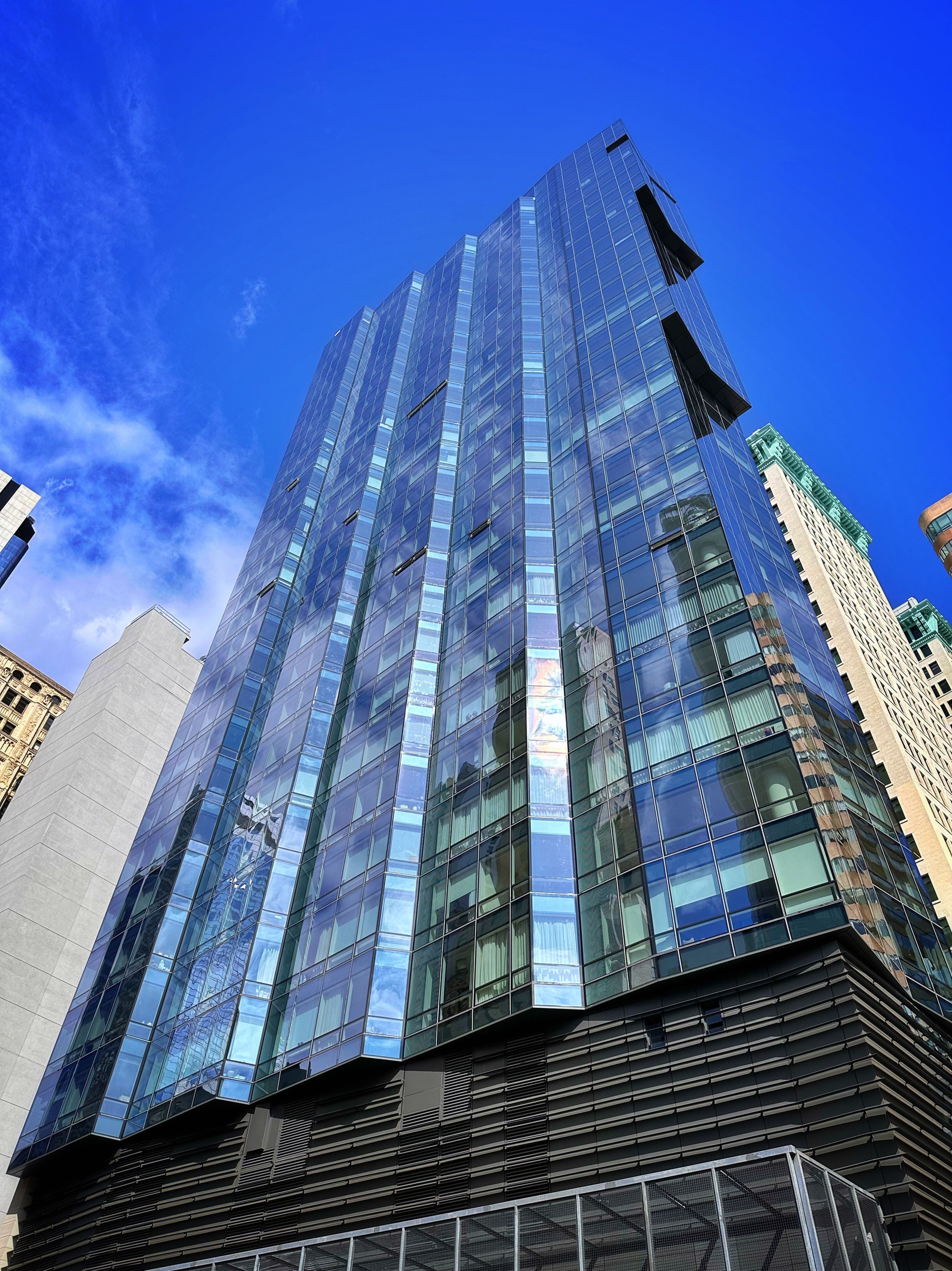 A tall modern glass skyscraper reflecting the blue sky, surrounded by other urban buildings under clear daylight.