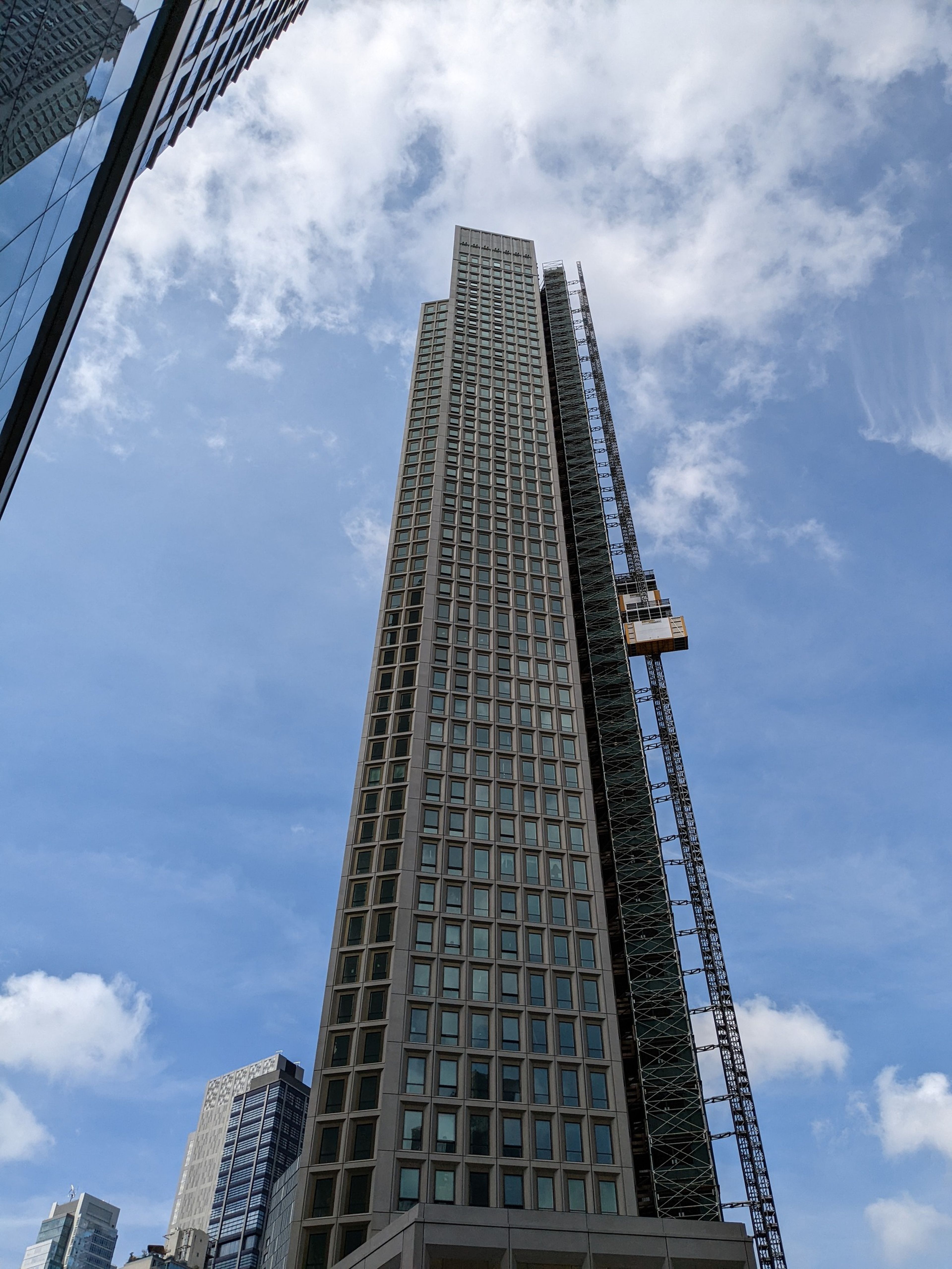 Tall skyscraper with grid-like windows reaching into a blue sky, flanked by smaller buildings. A construction lift is visible on the side.