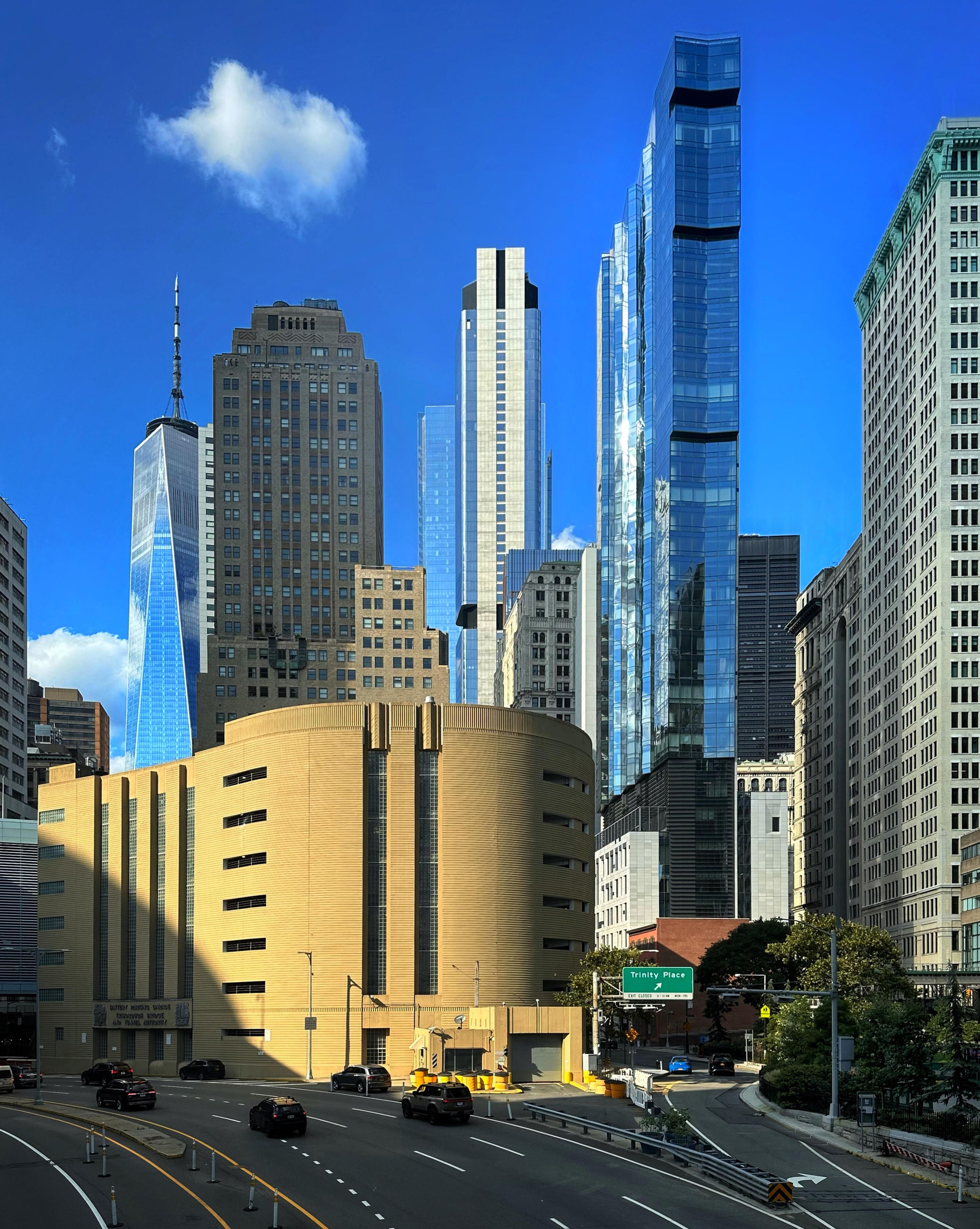 A cityscape featuring modern skyscrapers with reflective glass, a round tan building in the foreground, and a bright blue sky with a small cloud.