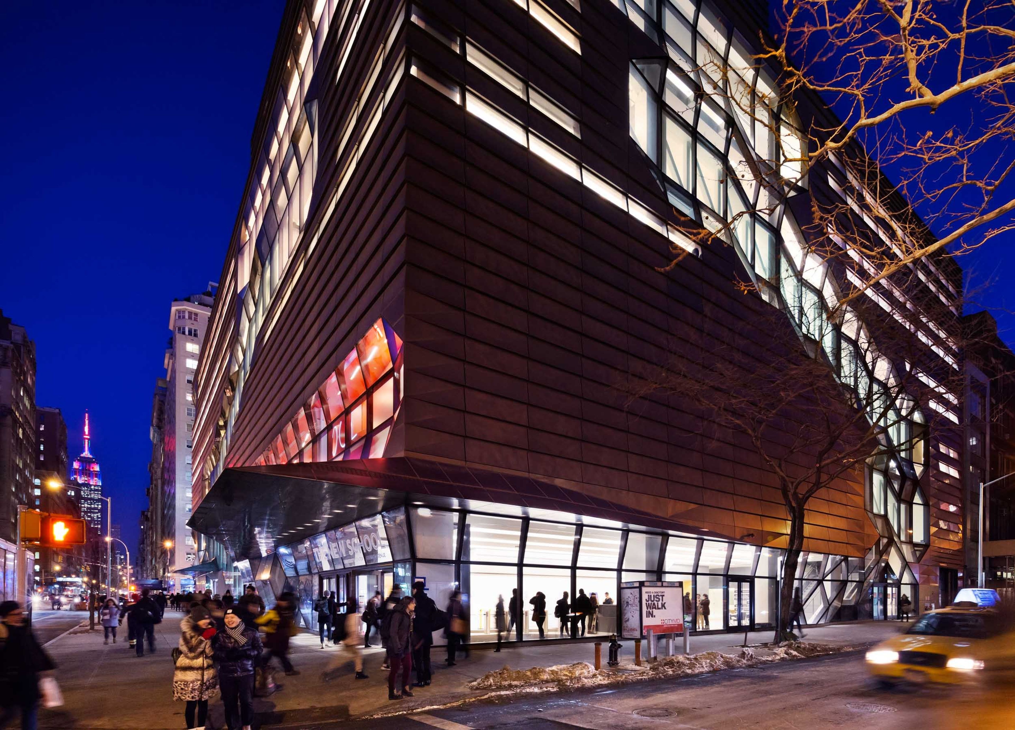 Modern building with angular design and illuminated windows at night, on a busy corner lot with many pedestrians passing in front. Cityscape with other buildings and traffic lights in the background and tree in the foreground. A yellow taxicab passes by in the street on the right.