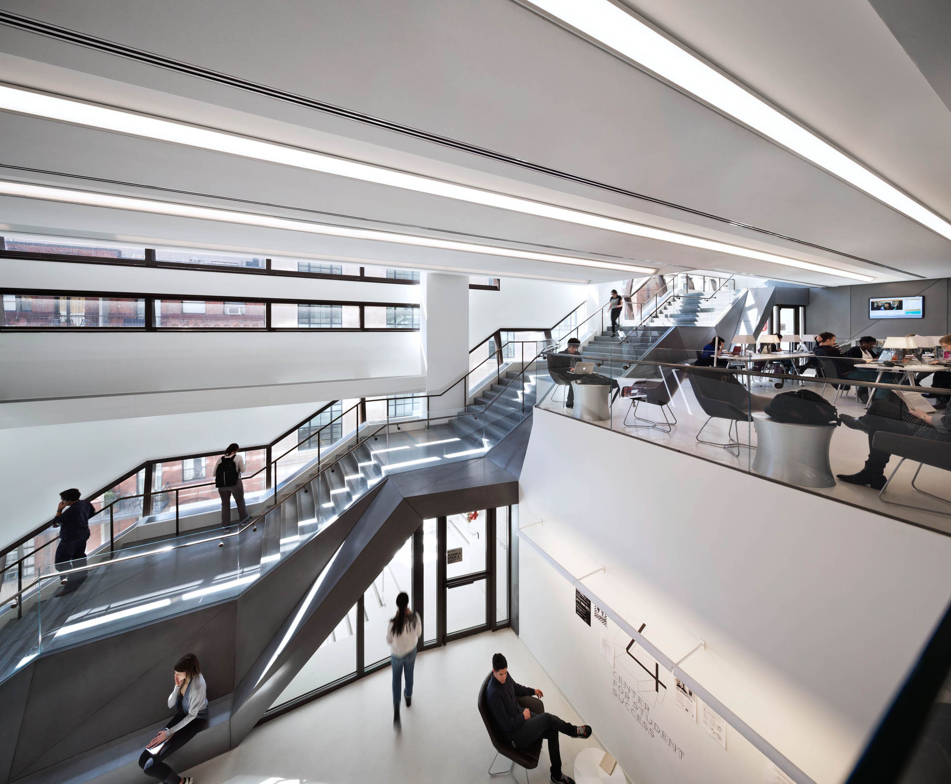 A bright, modern two-story entry space, featuring a large, continuous concrete stair with multiple landings in the background, in front of large windows. Gathering areas with people seated in the foreground.