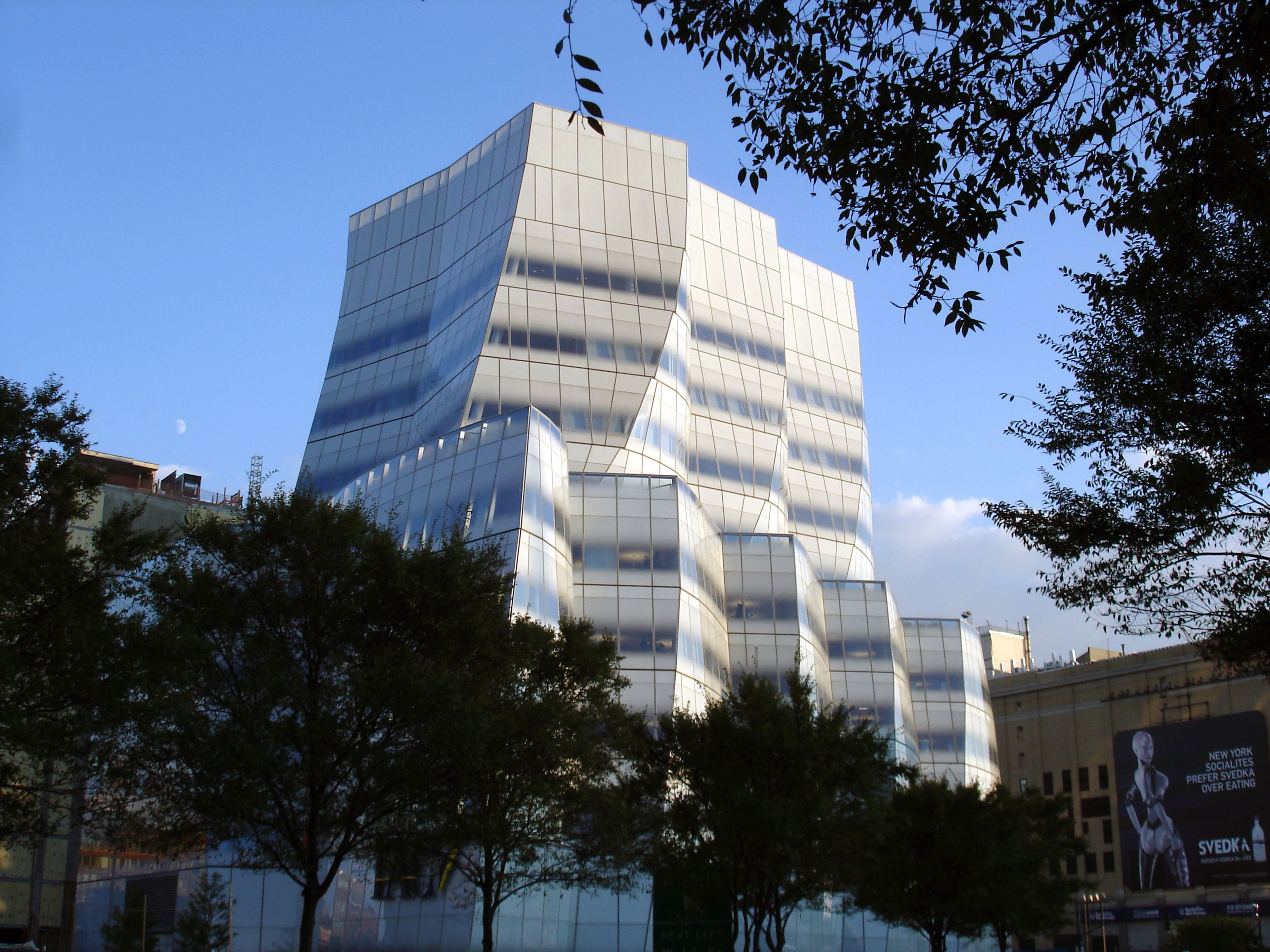 Modern, white building with a wavy, glass-and-metal facade against a clear blue sky, surrounded by trees and adjacent structures.