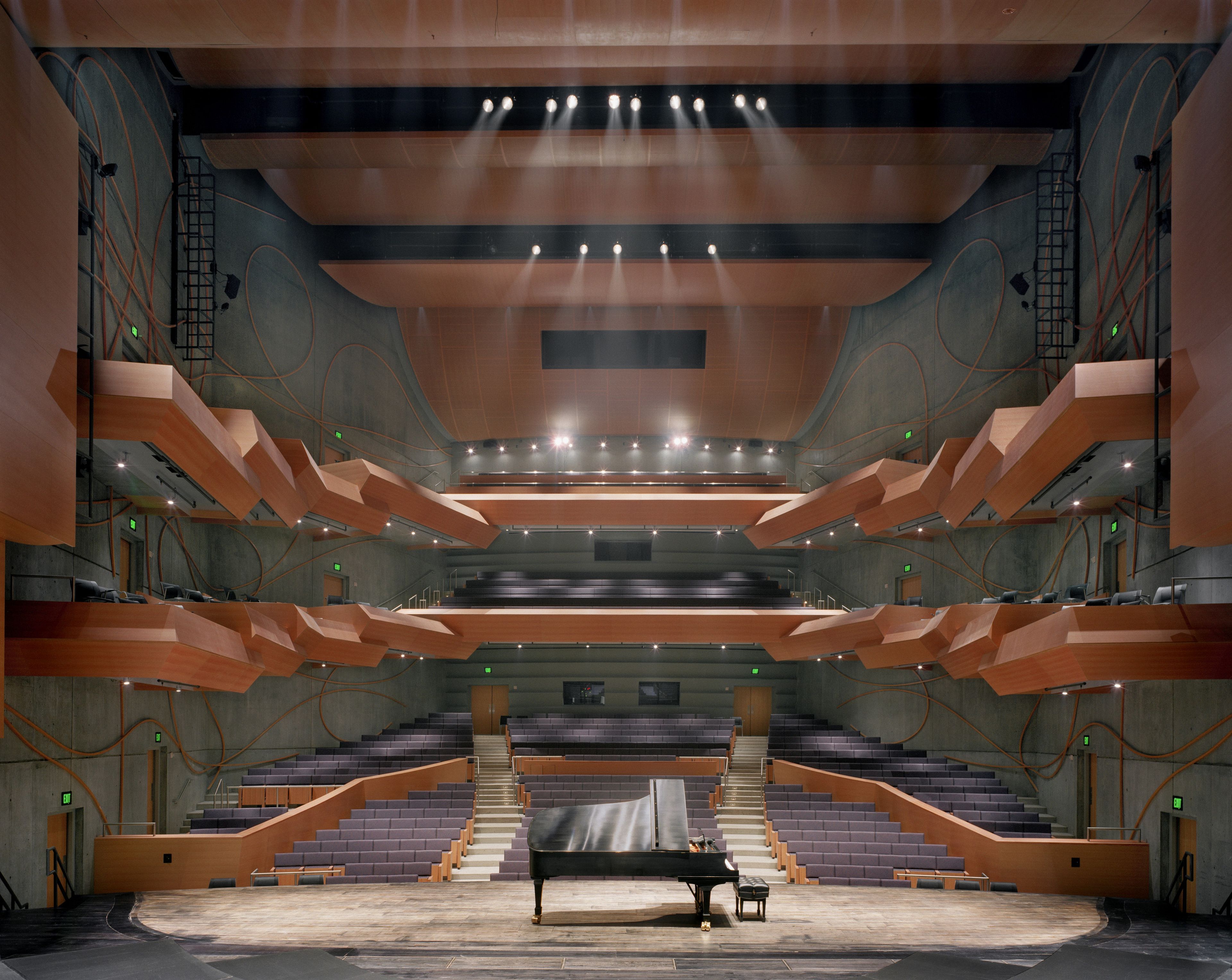 Modern concert hall interior with tiered seating, a grand piano on center stage, and dramatic lighting highlighting architectural details, like exposed concrete walls, plum colored seating, and warm wood paneling on the ceiling and balcony fronts..