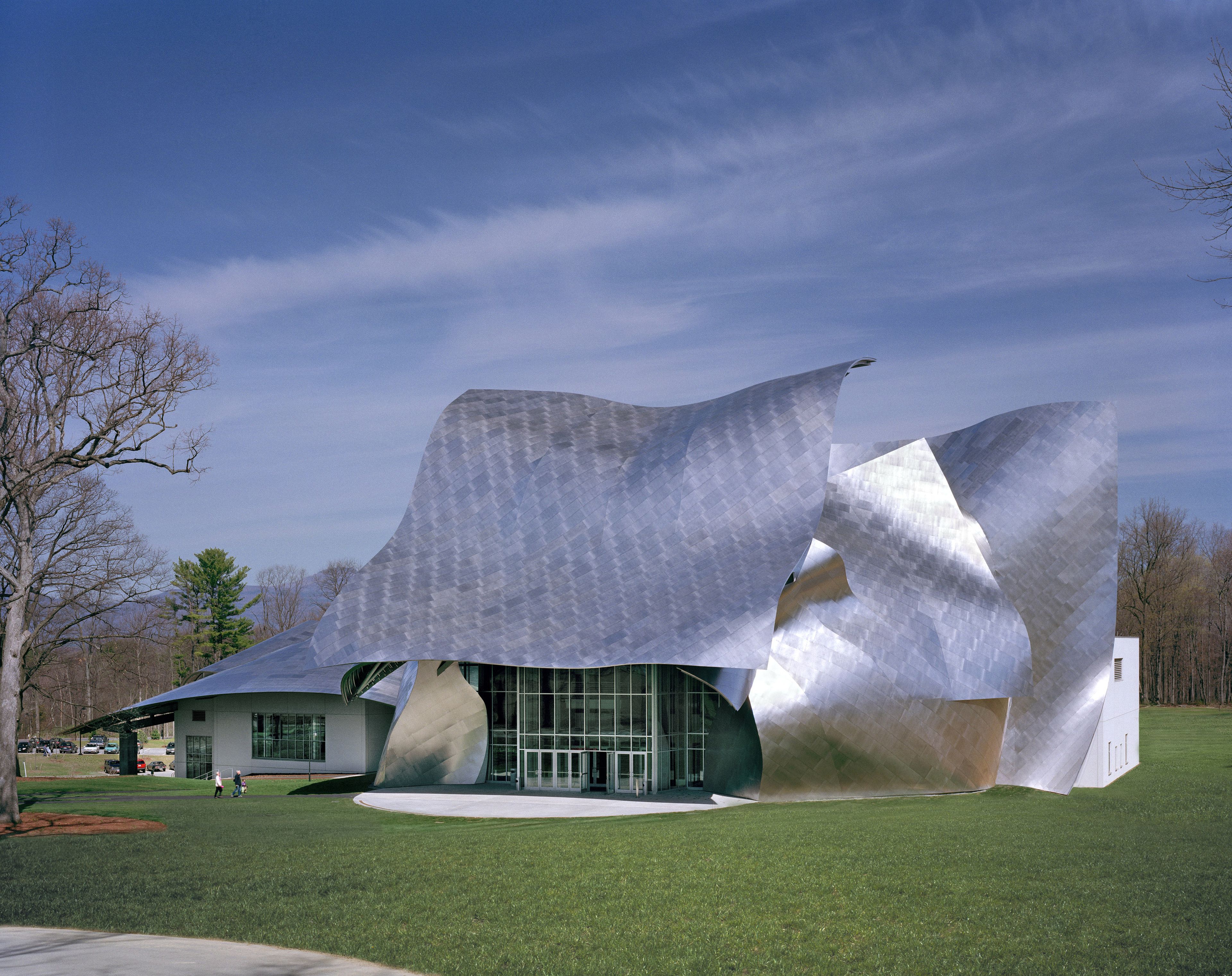 Modern building with a wavy, metallic exterior set against a blue sky with wispy clouds and surrounded by budding trees and a spacious, grassy lawn.