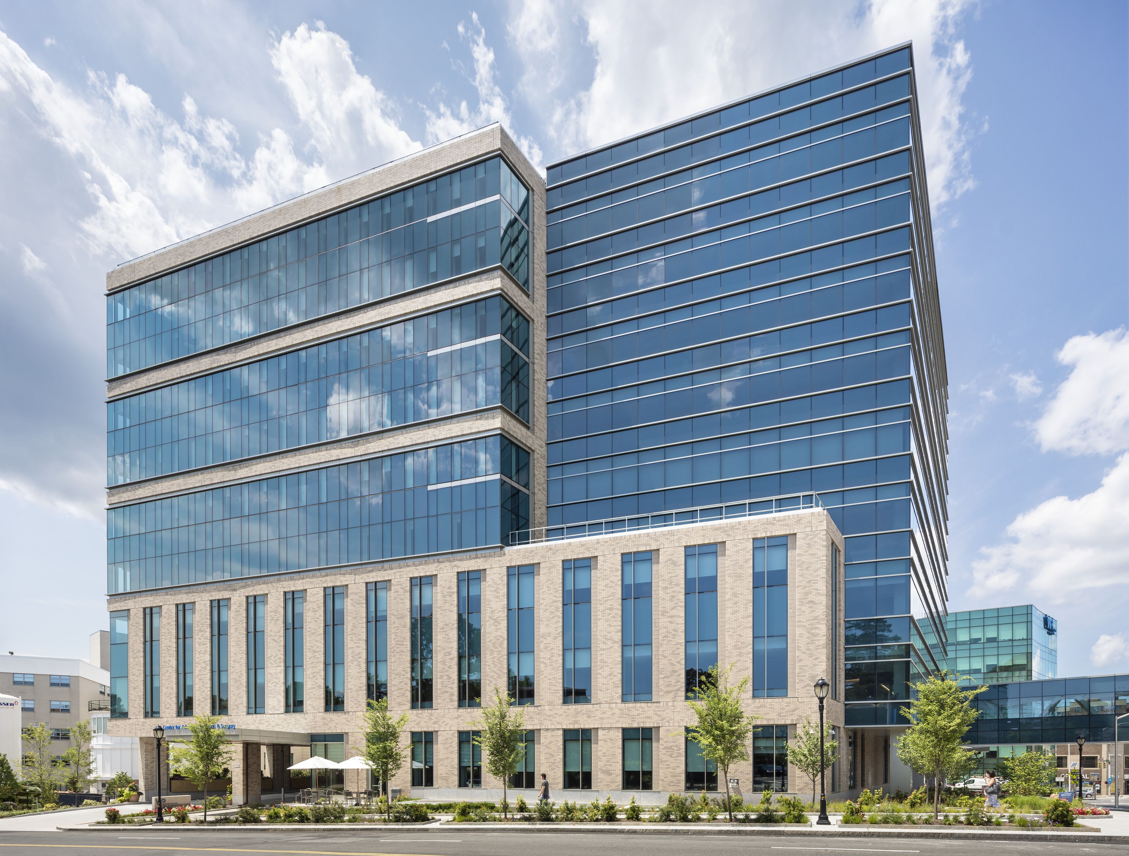 Modern medical building with glass and brick facade, multiple stories, and a landscaped entrance under a partly cloudy sky.