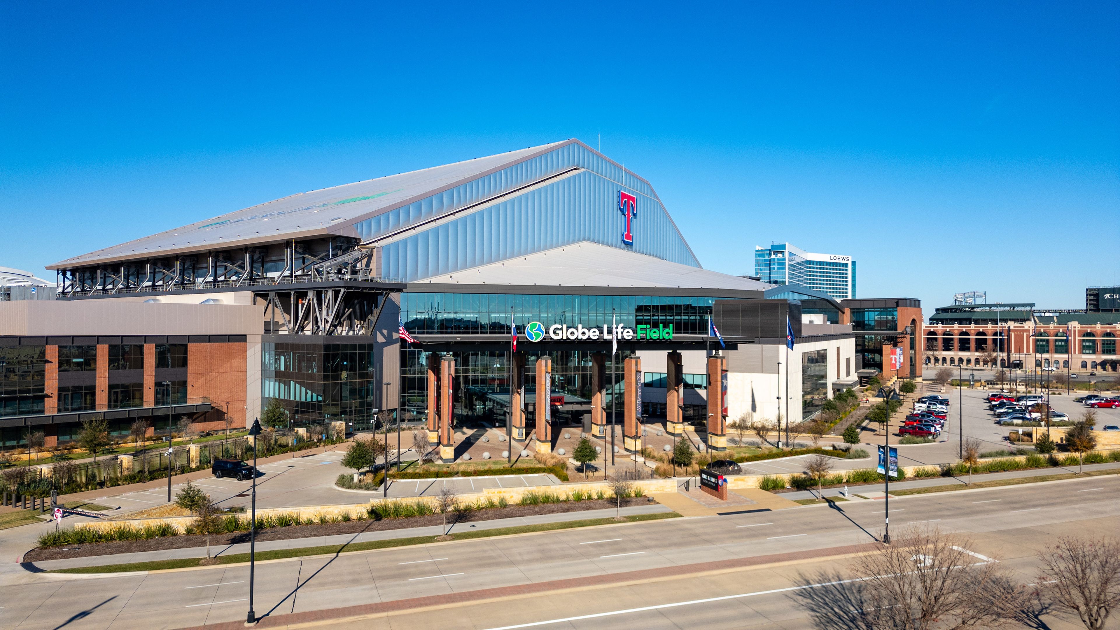 Exterior view of Globe Life Field, a modern stadium with a retractable roof, surrounded by parking, a road in the foreground, and a clear blue sky above.