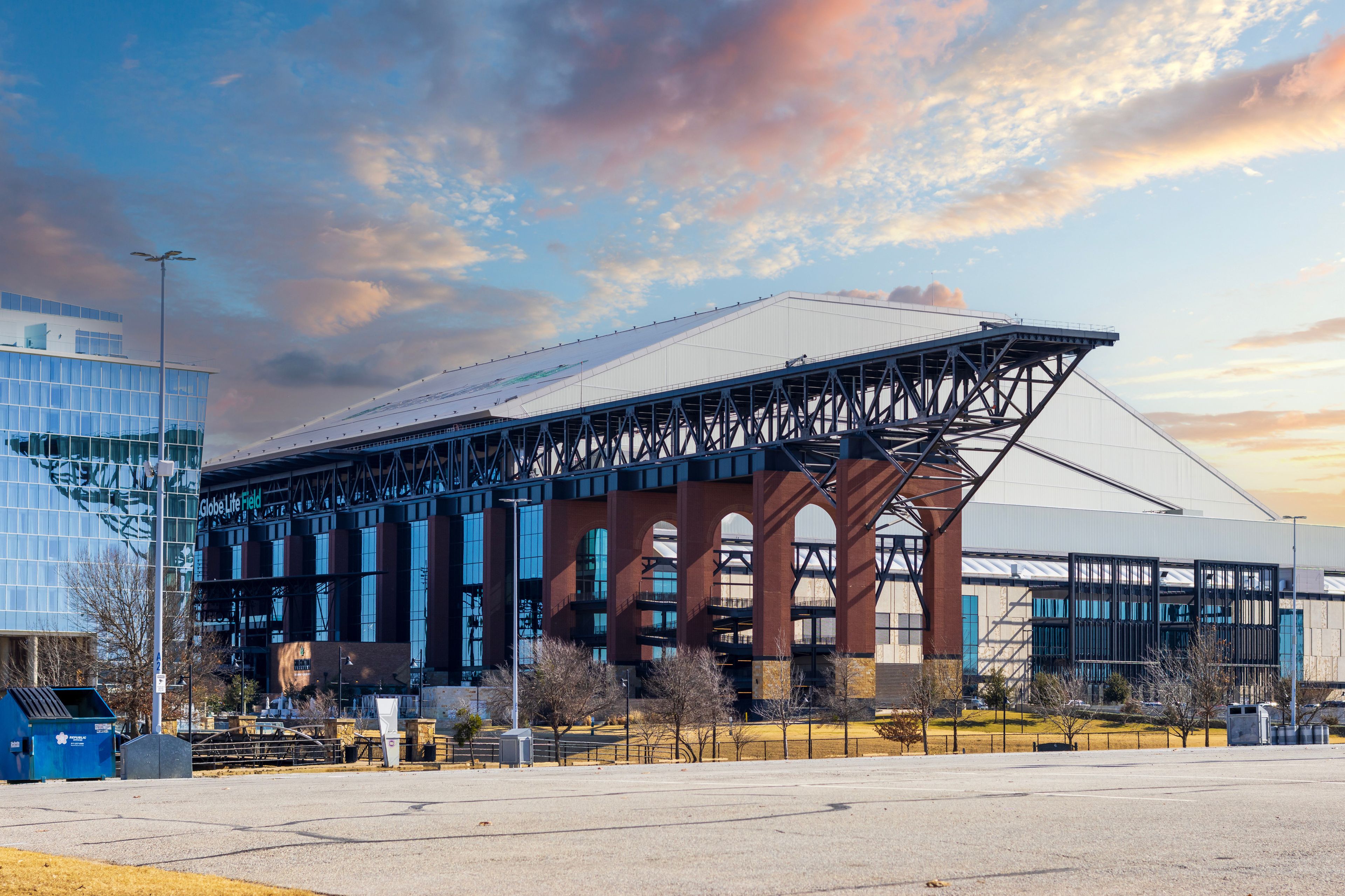 Set of large brick archways support retraction of adjacent stadium roof, under a colorful sunset sky. An empty parking lot and adjacent glass building are featured in the foreground