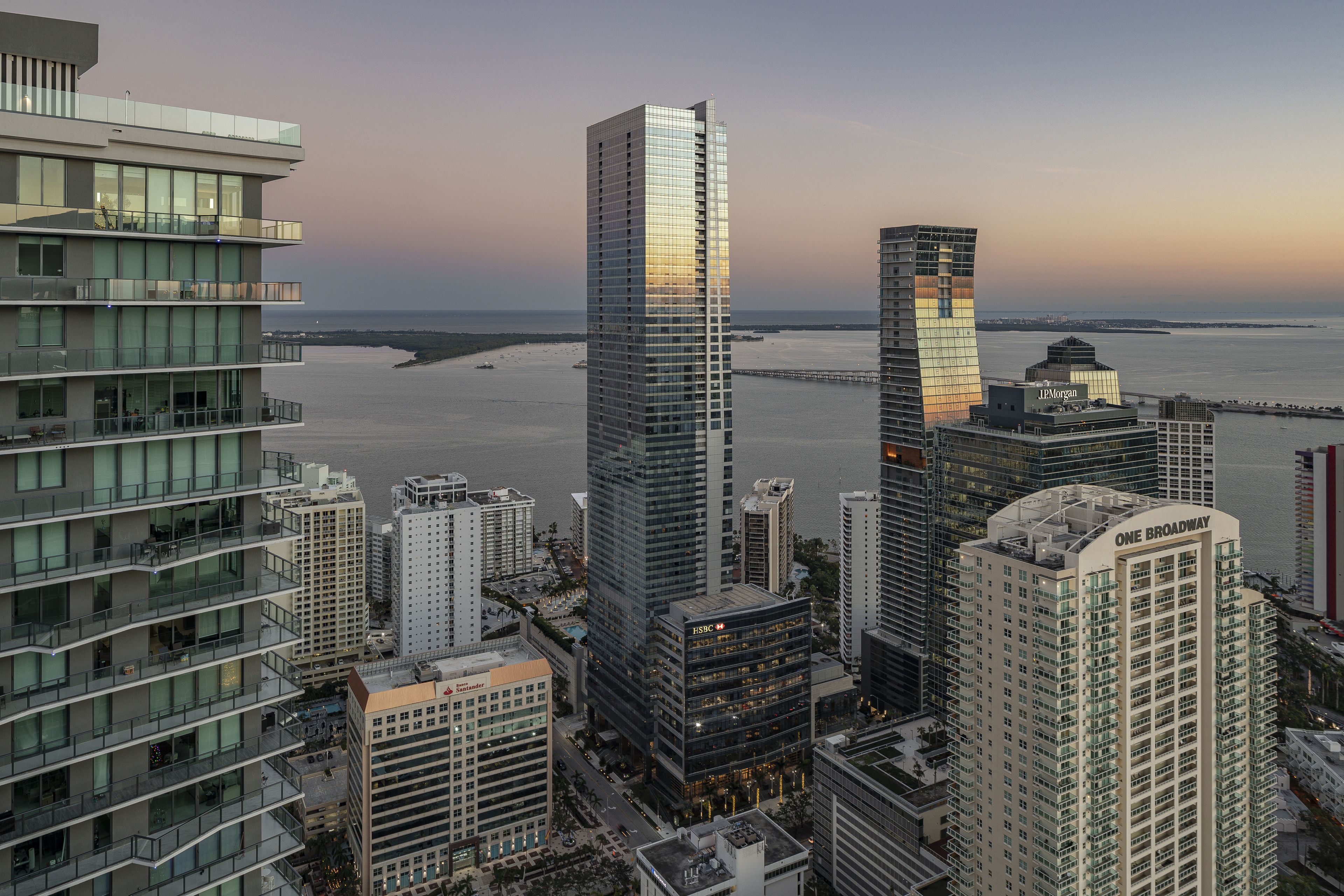Aerial view of a cityscape at sunset, featuring tall modern skyscrapers and a bay in the background.