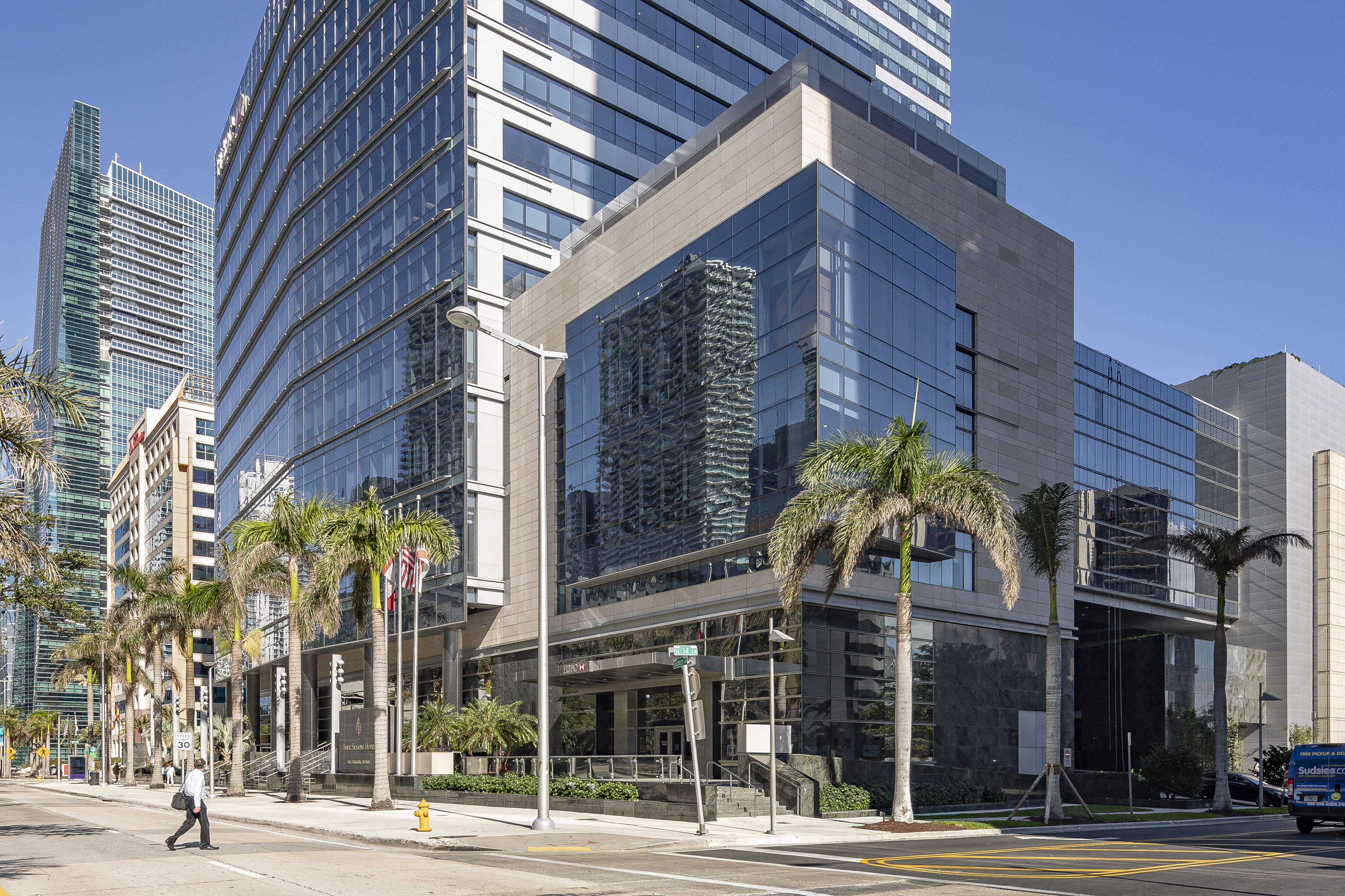 Street corner with modern glass and stone building in foreground and taller glass buildings in the background under a clear blue sky. A person crosses the palm-tree lined street in the foreground.