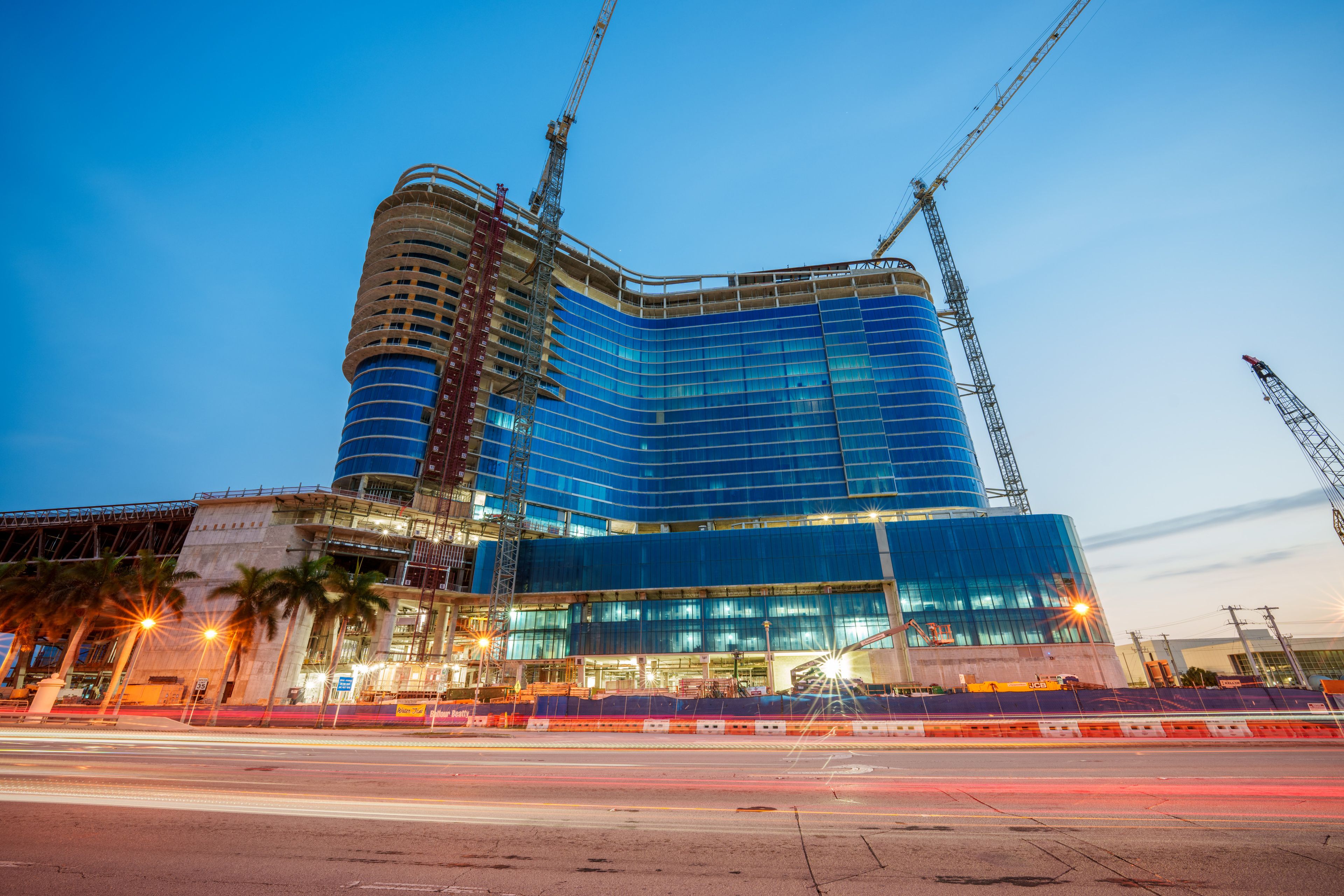 Construction site of a modern multi-story building with blue glass facade, cranes, and palm trees at dusk, with light trails from passing vehicles.