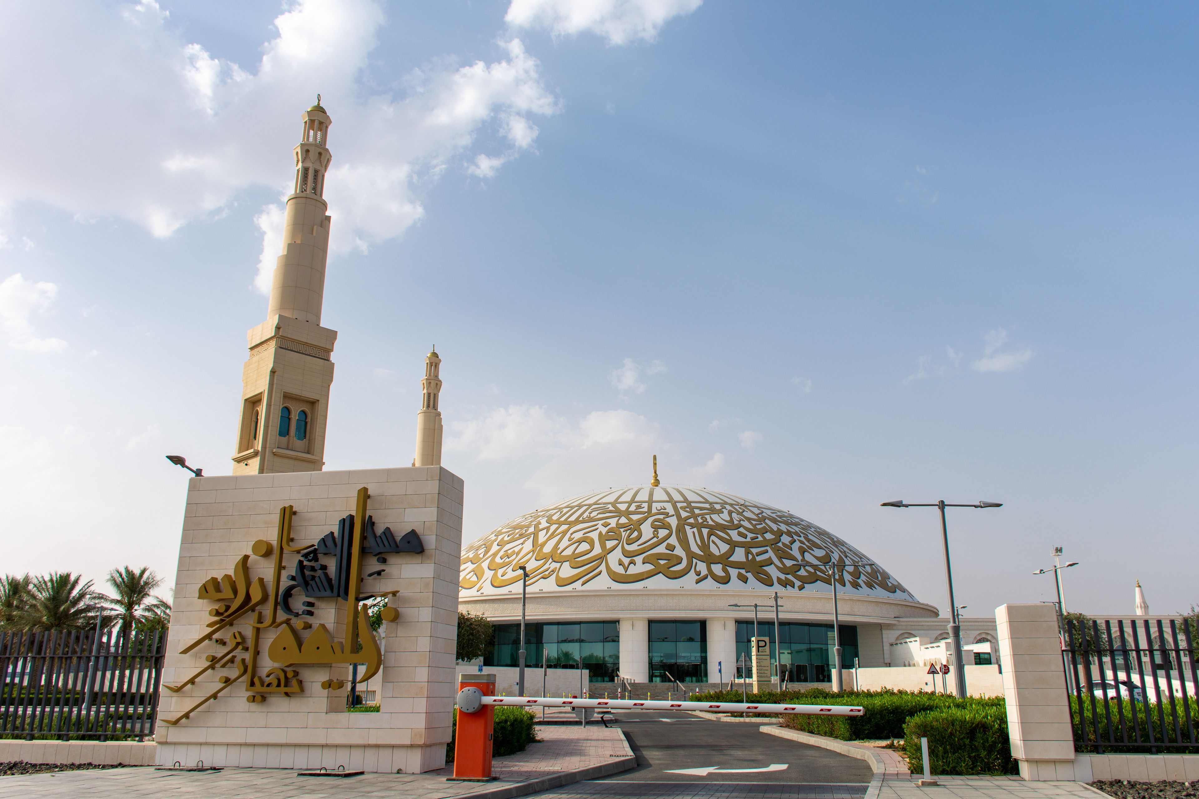 A mosque with a large dome featuring intricate Arabic calligraphy, flanked by tall minarets and surrounded by palm trees under a blue sky.