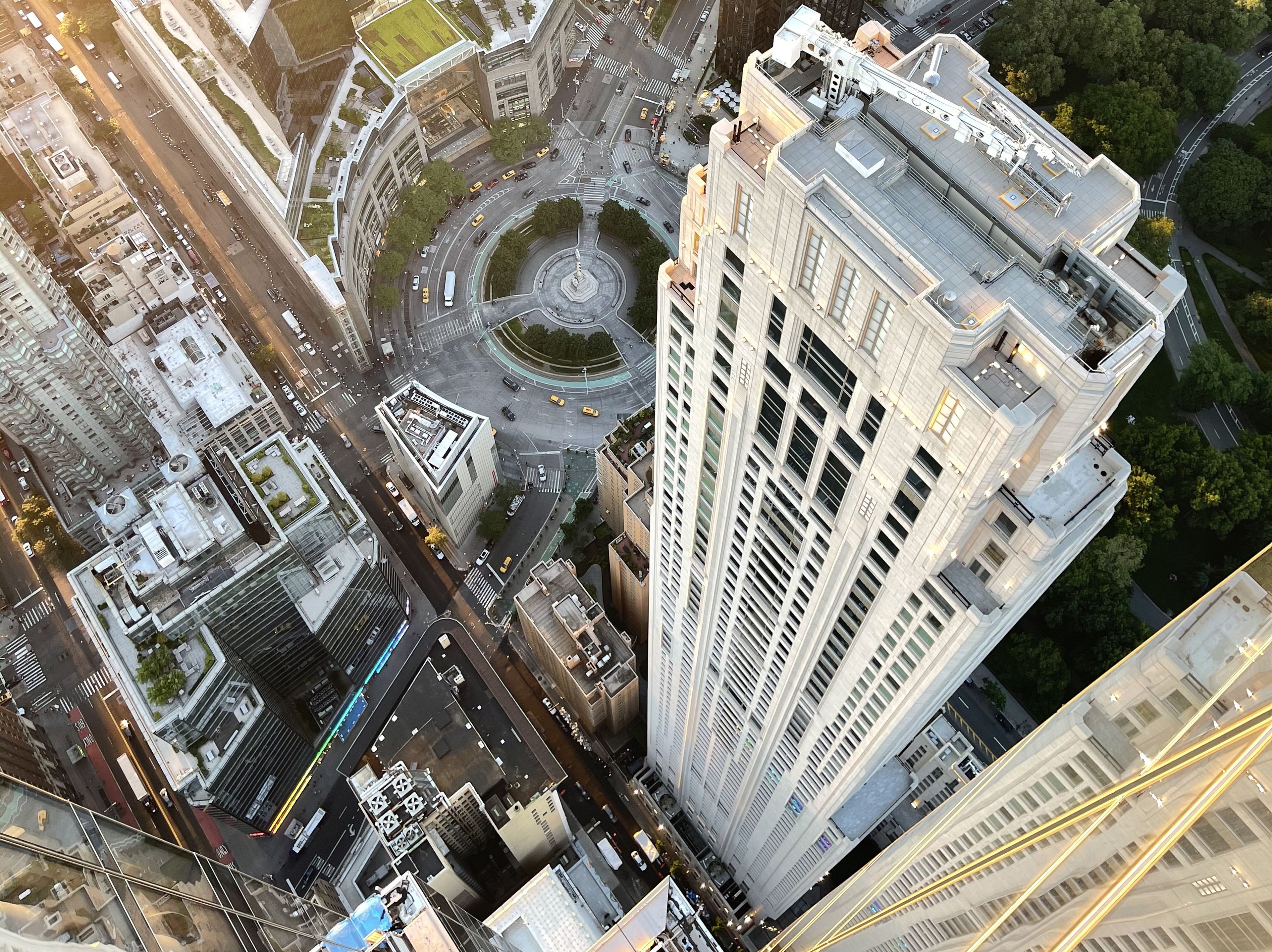 Aerial view of a tall skyscraper in a cityscape, surrounded by streets, shorter buildings, and a roundabout with trees and a fountain. A larger park is visible behind the building.