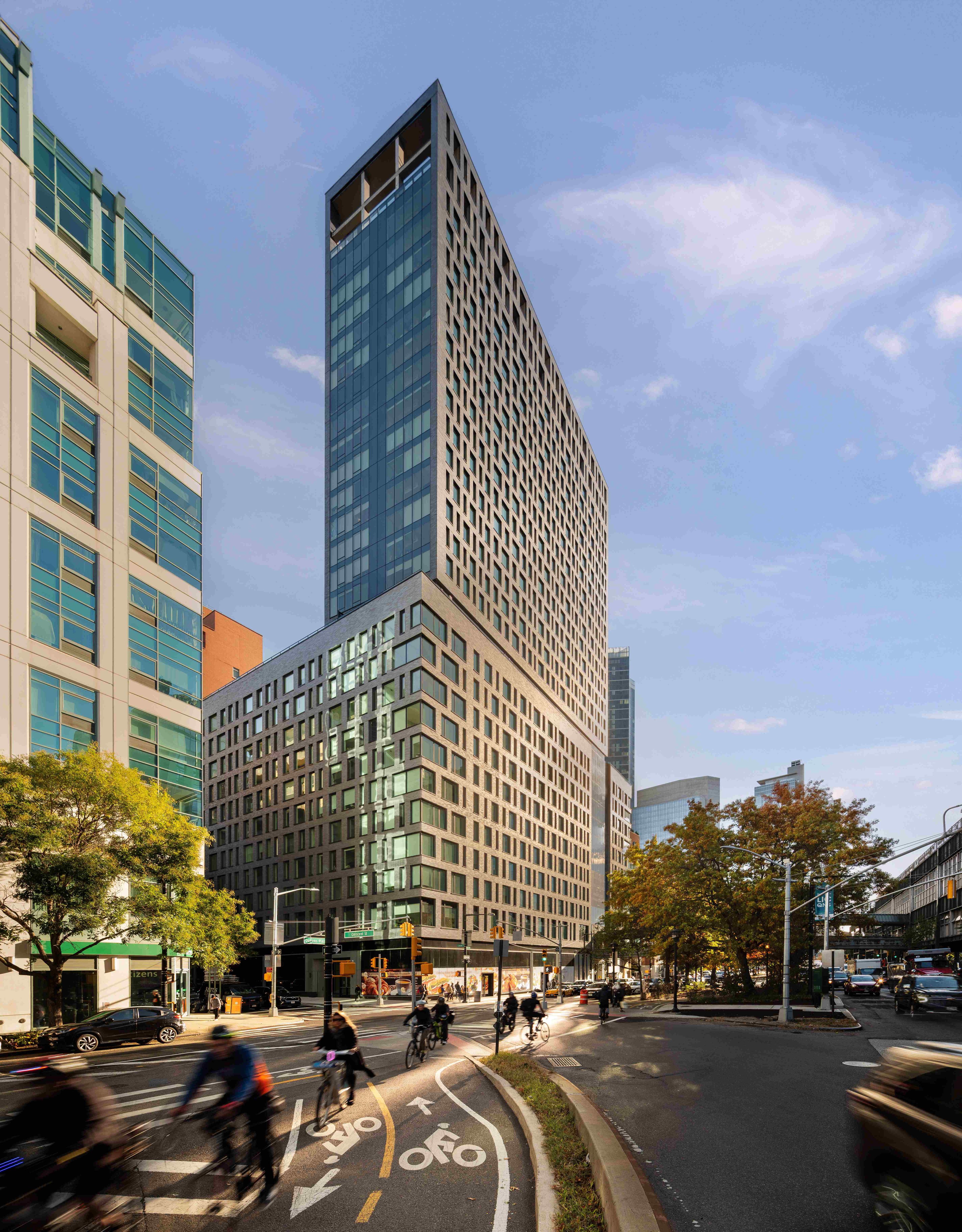 Tall modern building with glass windows on a bustling city street, surrounded by cyclists and trees, under a clear blue sky.
