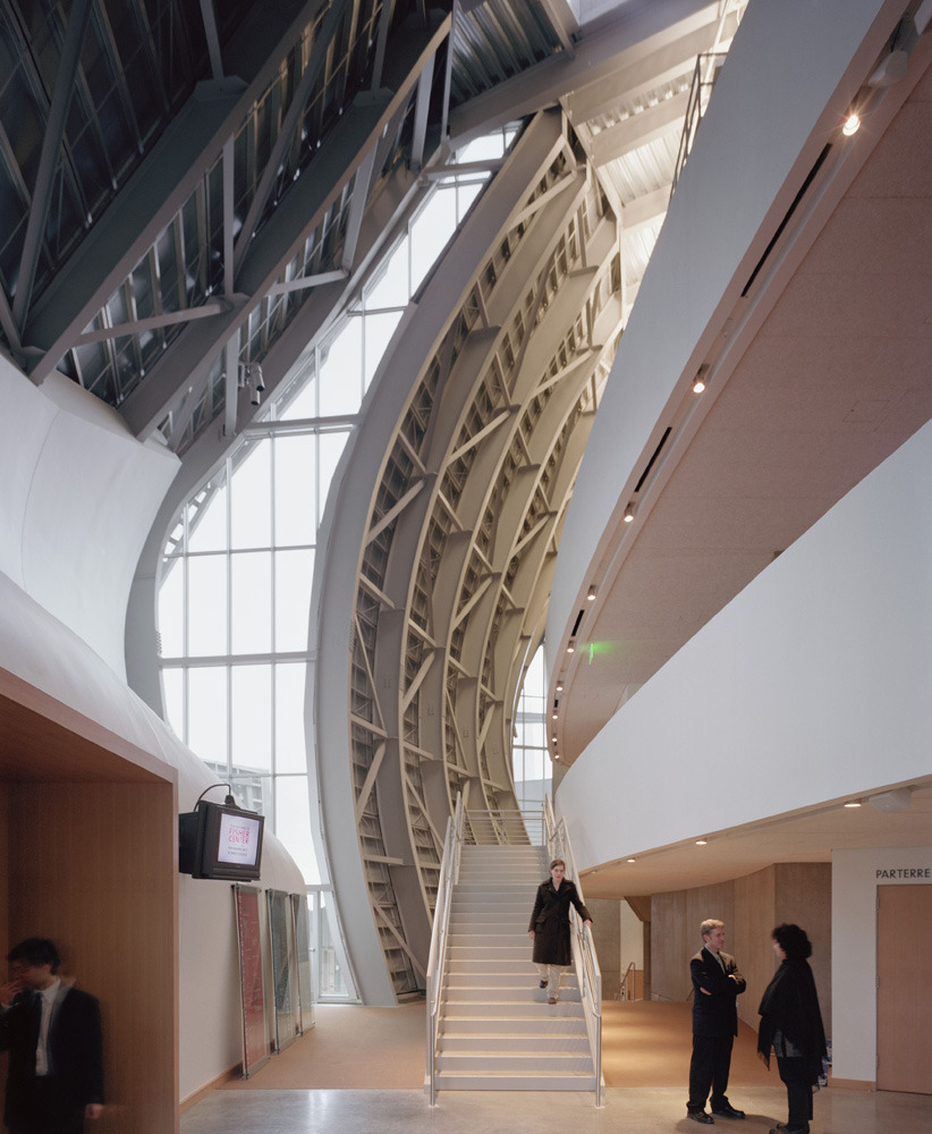 Modern atrium interior with a curved glass-and-metal exterior wall with exposed structural cross-bracing. In the center of the image is a single-story staircase with a person standing on it, midway down and another couple of people standing at its base conversing..