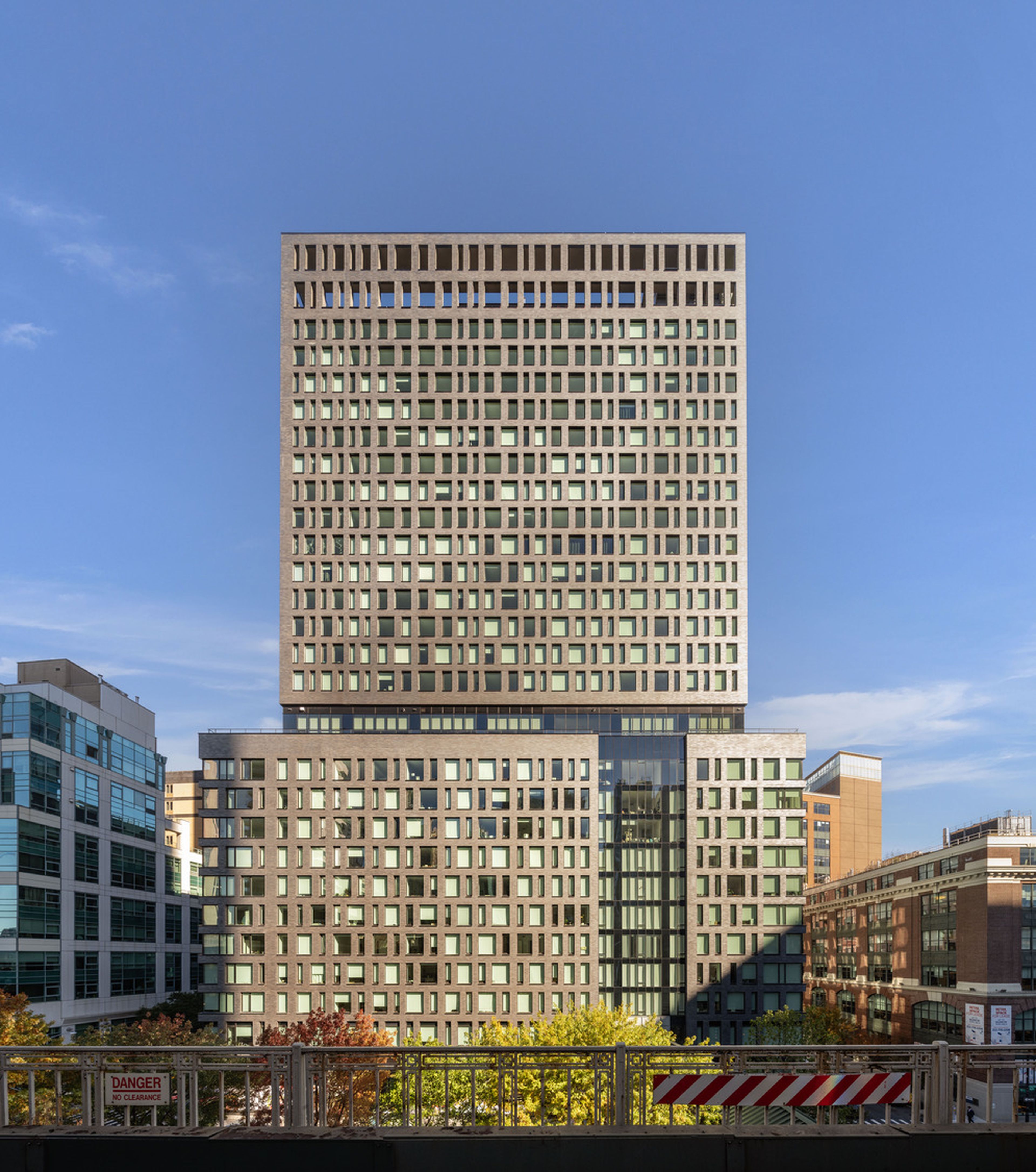 Tall modern building with a grid-like window pattern, against a blue sky, surrounded by smaller buildings and trees in an urban setting.