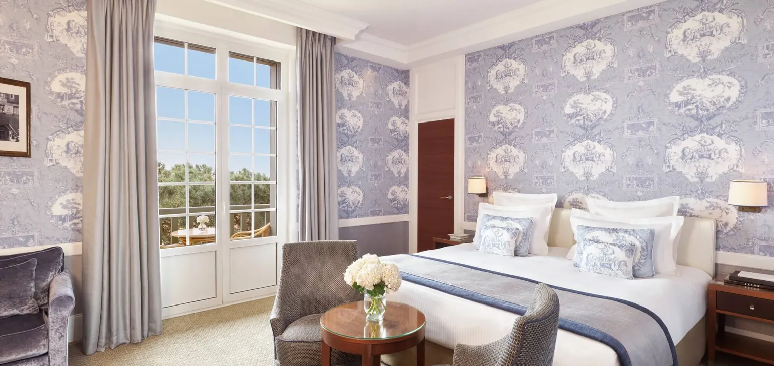 Chambre d'hôtel élégante avec un lit double, un papier peint à motifs bleus, des fauteuils, une petite table ronde avec des fleurs et une grande fenêtre avec une vue panoramique.
