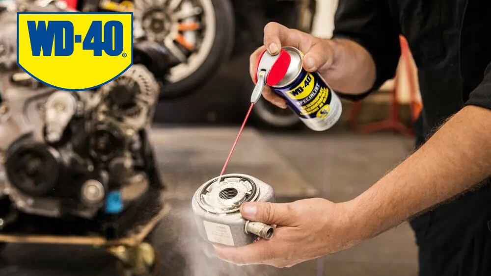 Mechaniker sprüht WD-40 Specialist auf ein Kfz-Bauteil in einer Werkstatt, im Hintergrund ein Automotor.