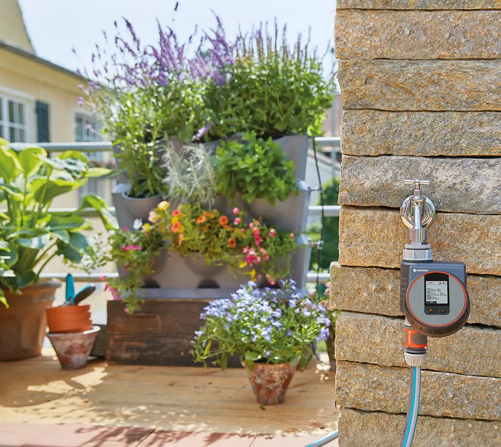 Gardena Bewässerungscomputer an Steinmauer mit Wasserhahn, im Hintergrund bepflanzter Balkon mit bunten Blumen und Kräutern.