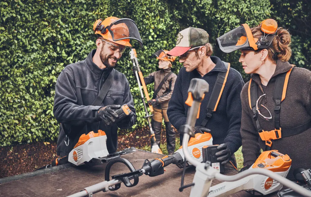 Drei Personen mit STIHL-Schutzhelmen begutachten Akku-Gartengeräte auf einem Tisch, im Hintergrund arbeitet eine weitere Person.