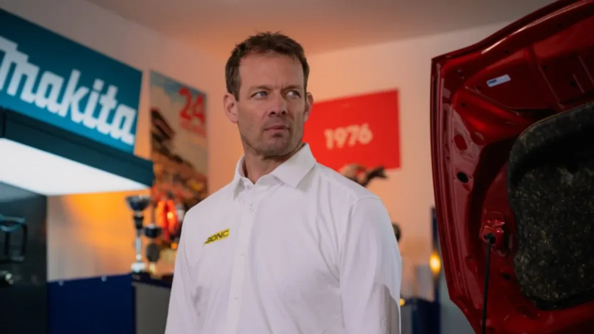 Mann in weißem Hemd mit Logo steht in einer Autowerkstatt neben einem roten Auto, Makita-Schild im Hintergrund.